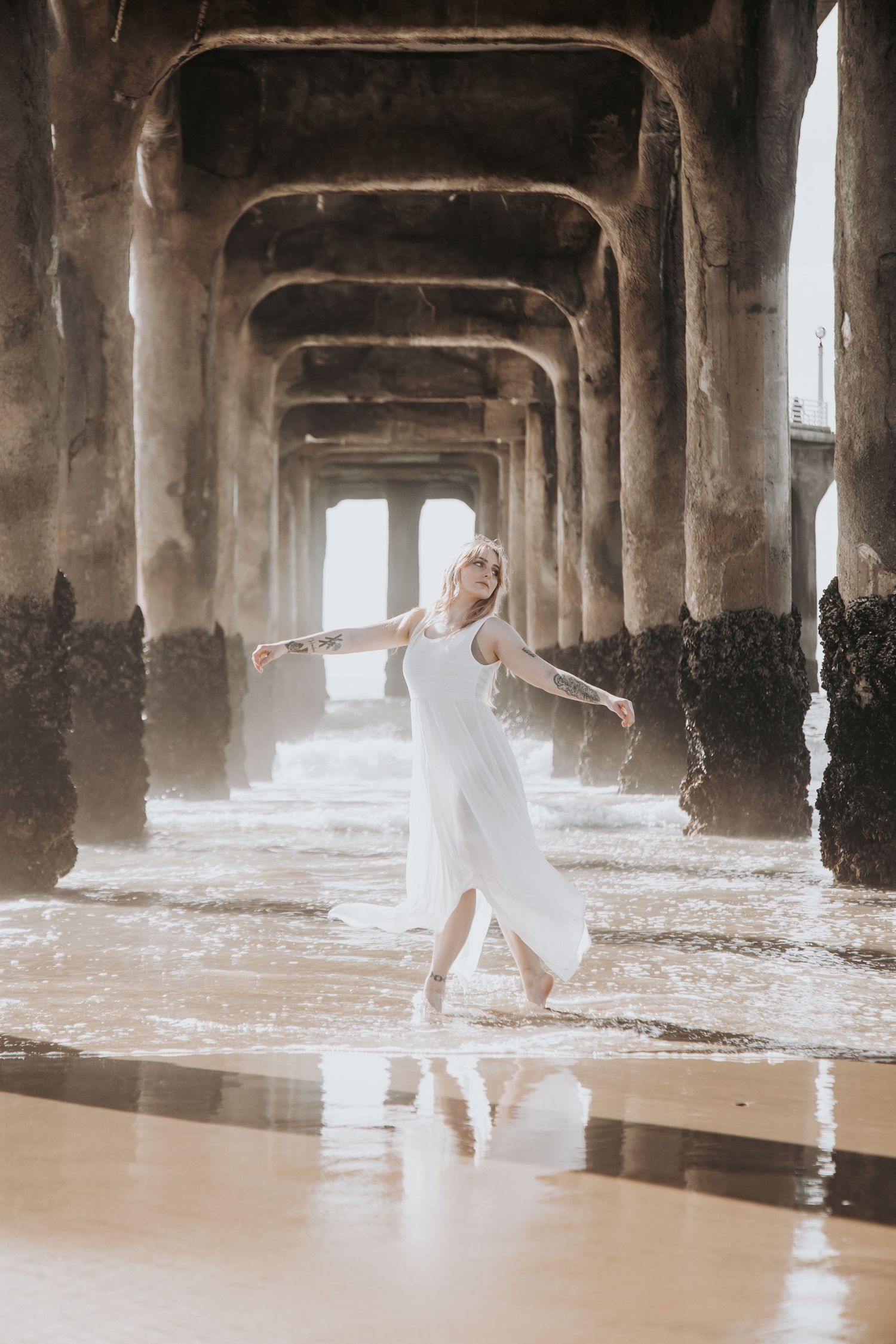 Woman in flowing white dress dancing gracefully beneath weathered pier pillars with ocean waves around her.
