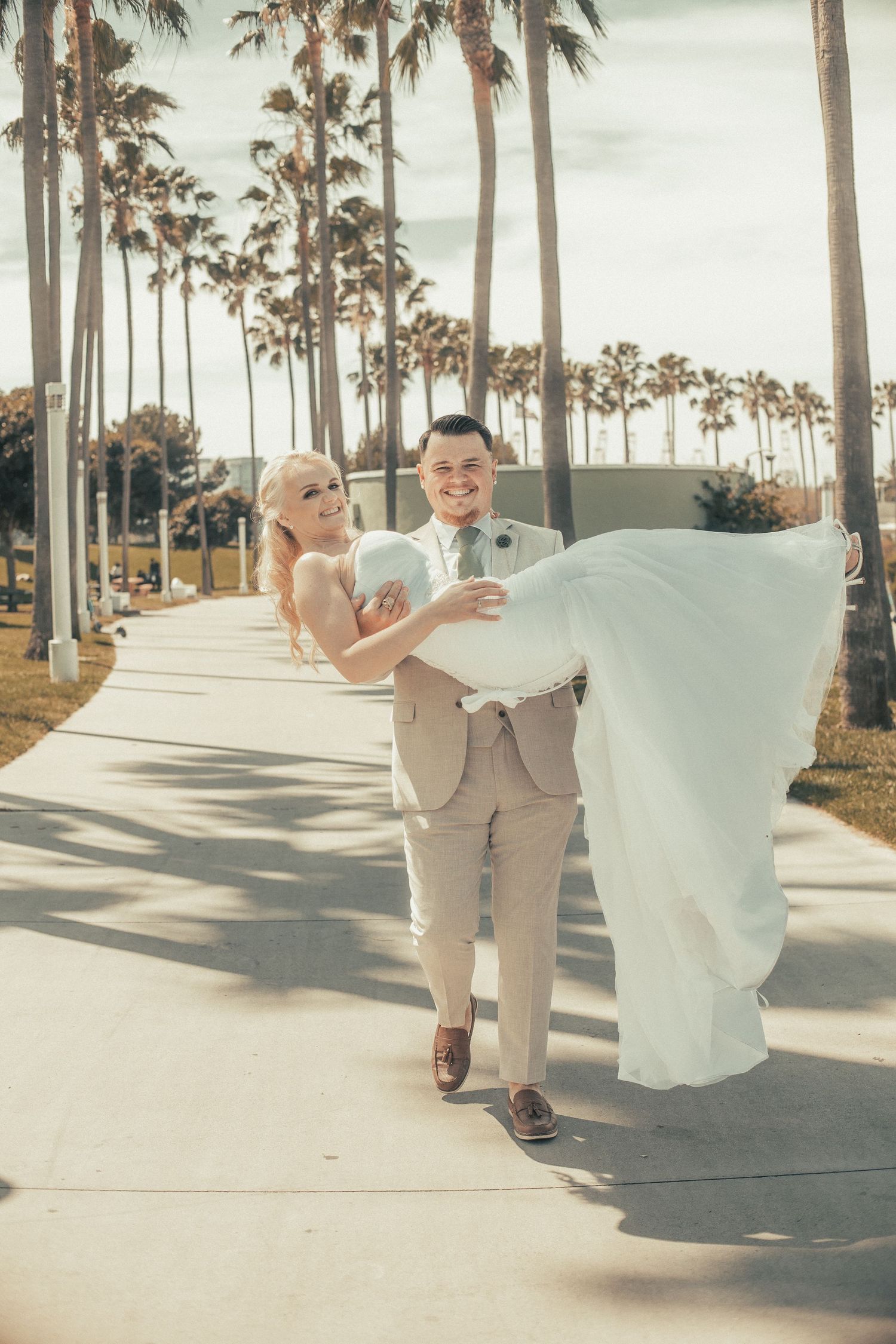 Groom in tan suit carries bride in flowing white dress along palm tree-lined walkway.