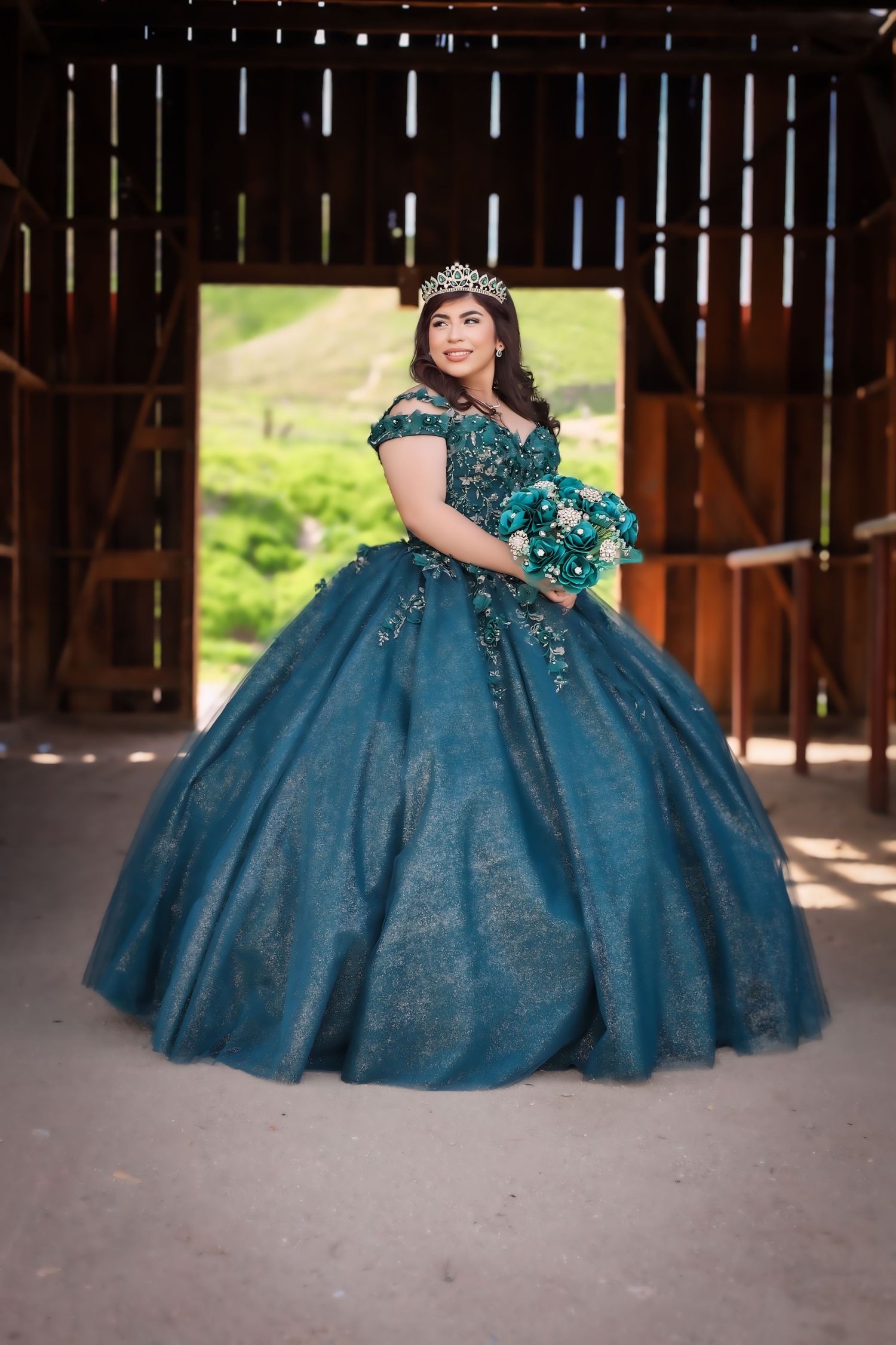 Young woman in teal quinceañera ball gown with tiara holds bouquet inside rustic barn.