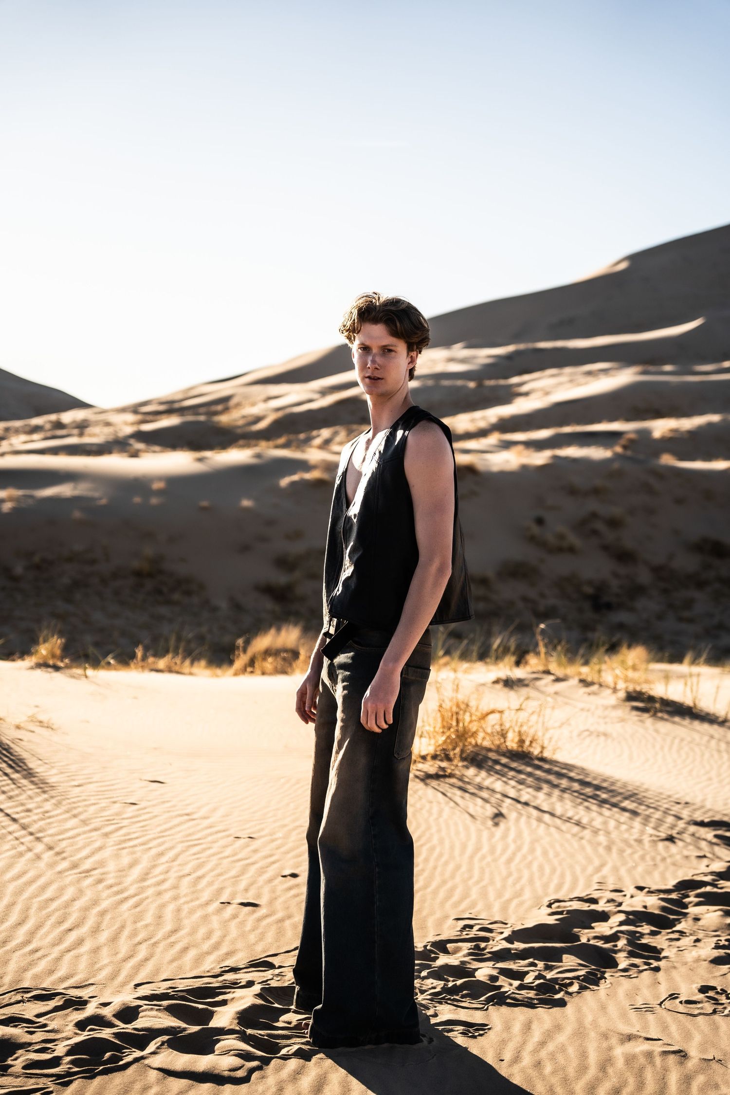 Young man in black sleeveless outfit standing on sandy desert dunes with large dunes in background.