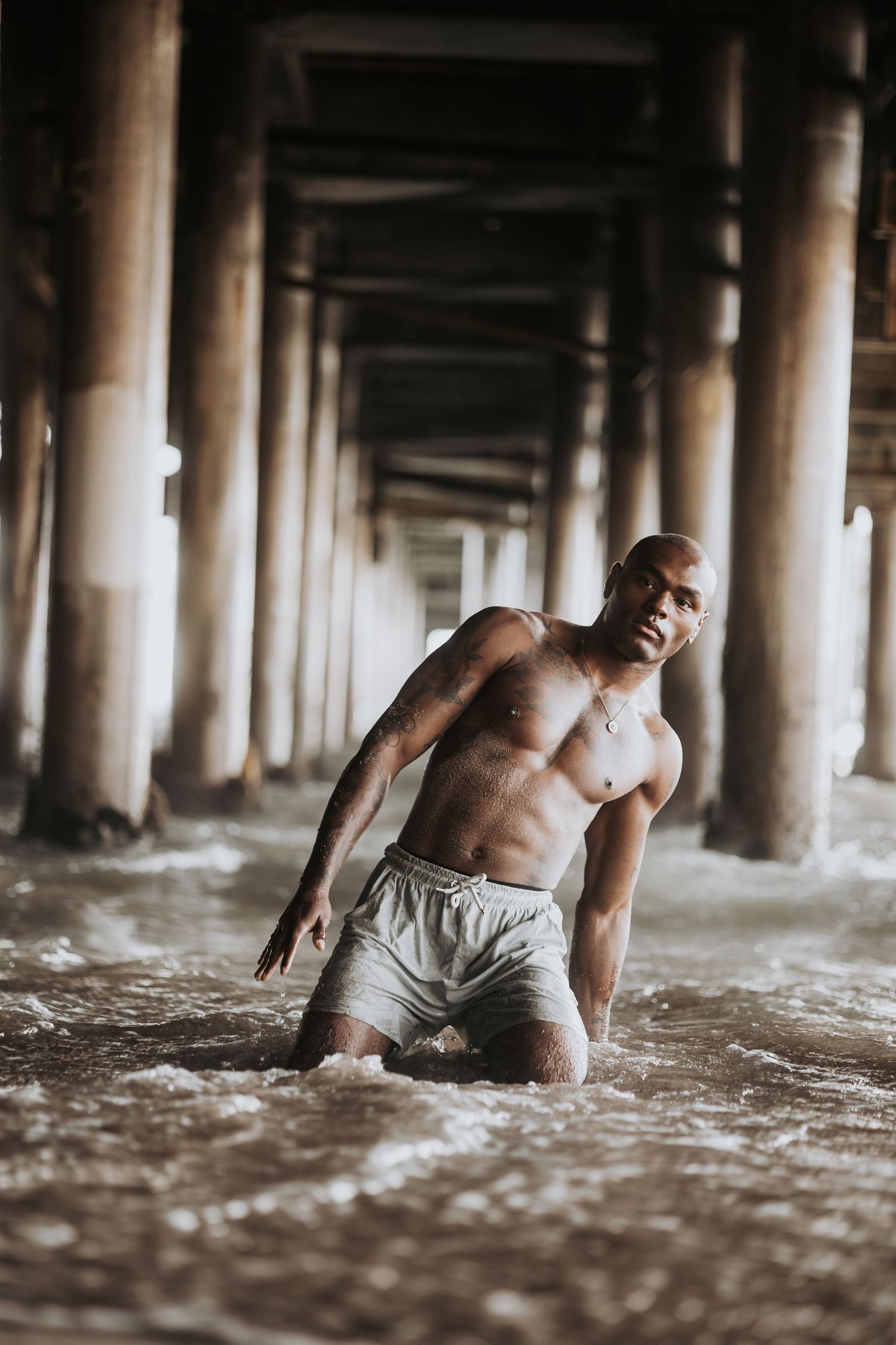 A shirtless muscular man kneels dramatically in shallow water beneath a beach pier.