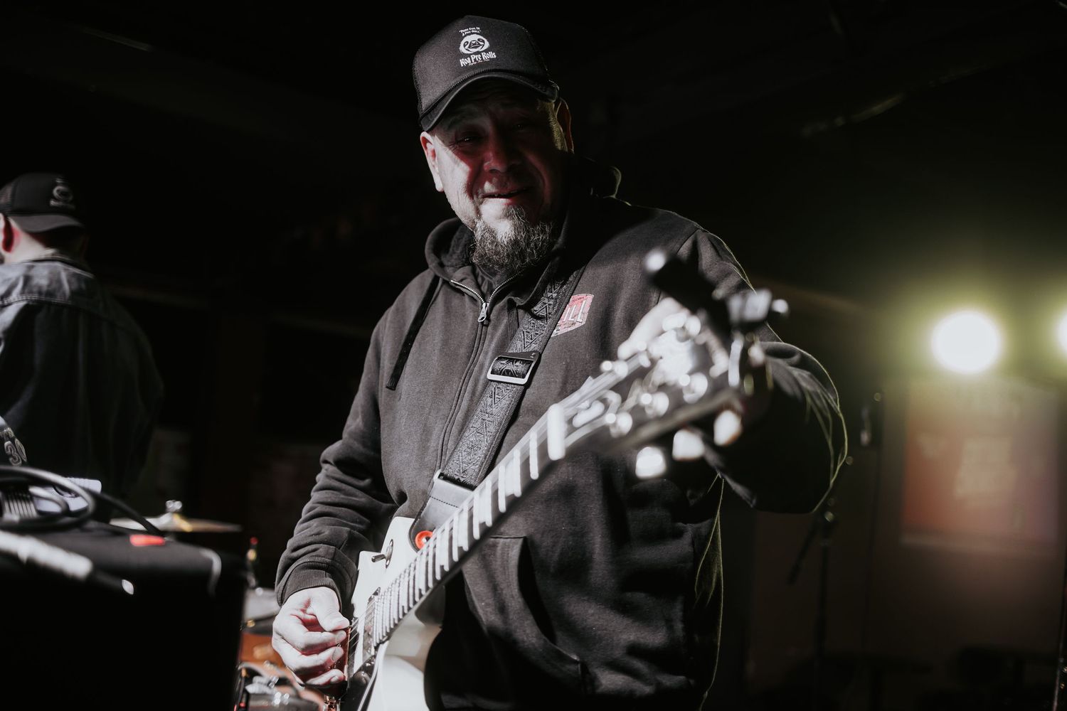 Male guitarist smiling while playing electric guitar on a dark stage wearing a black hoodie and cap.