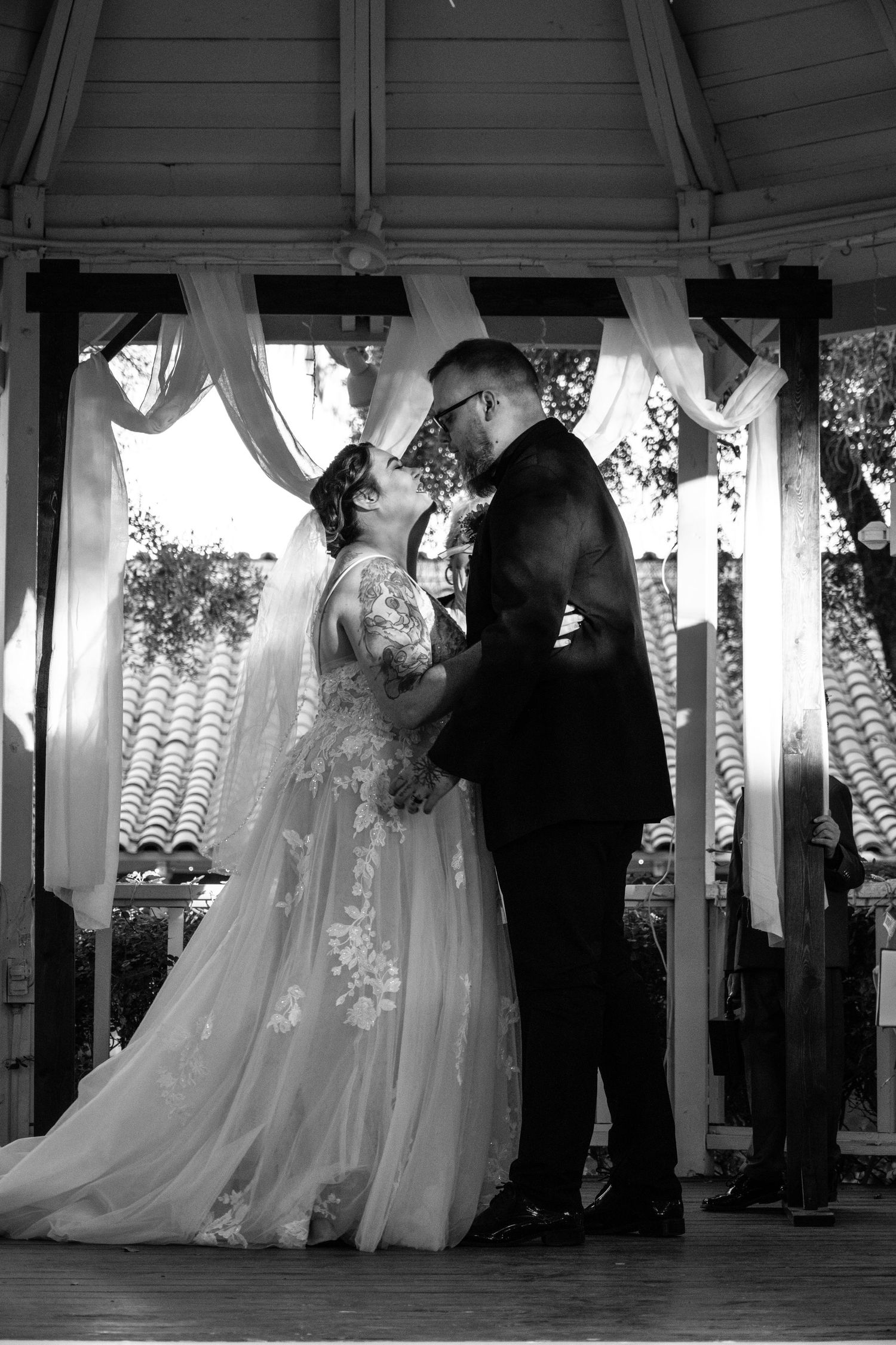 Bride and groom sharing first kiss under a draped gazebo in black and white wedding photo.