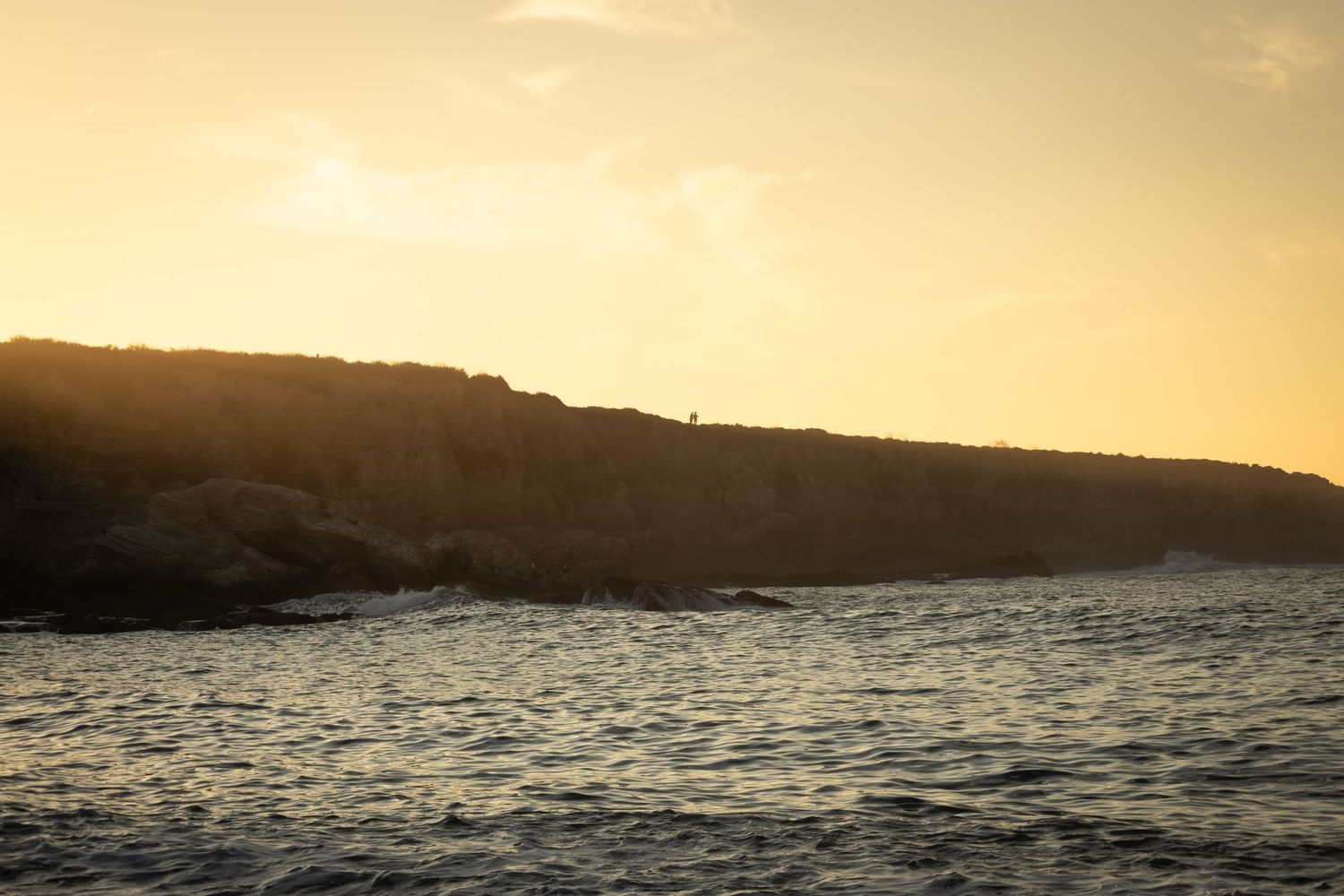 Golden sunset over rocky coastal cliffs with calm ocean waves in the foreground.