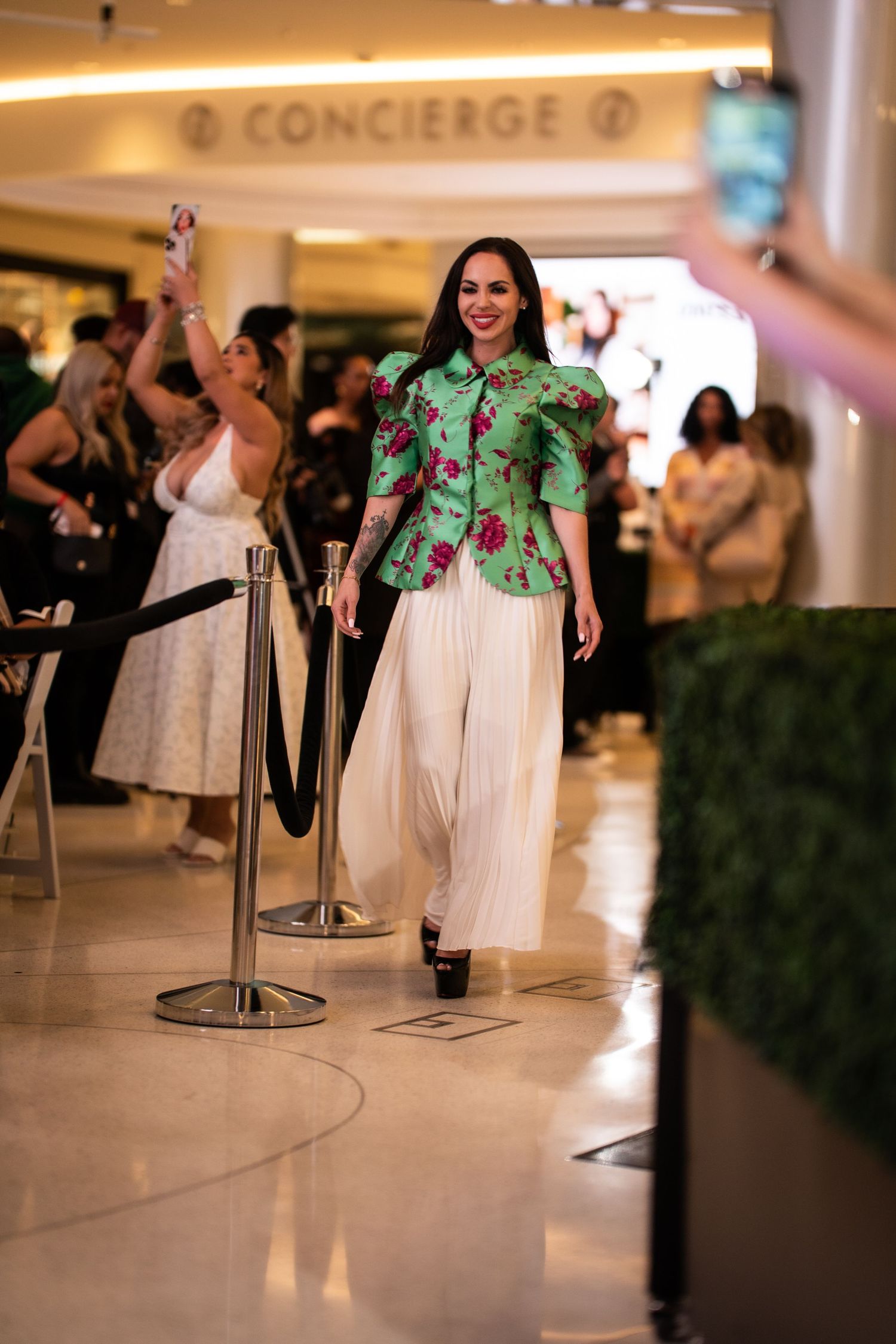 A woman walks a runway in a green floral peplum top and white wide-leg pants.