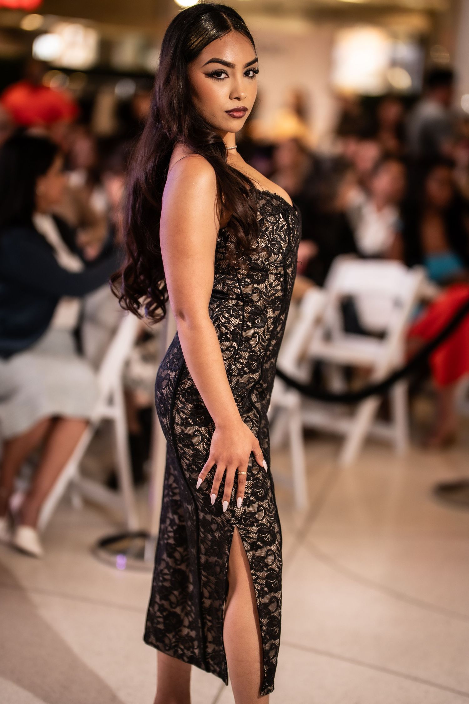 A woman poses in a black lace midi dress with a slit at a fashion show event.