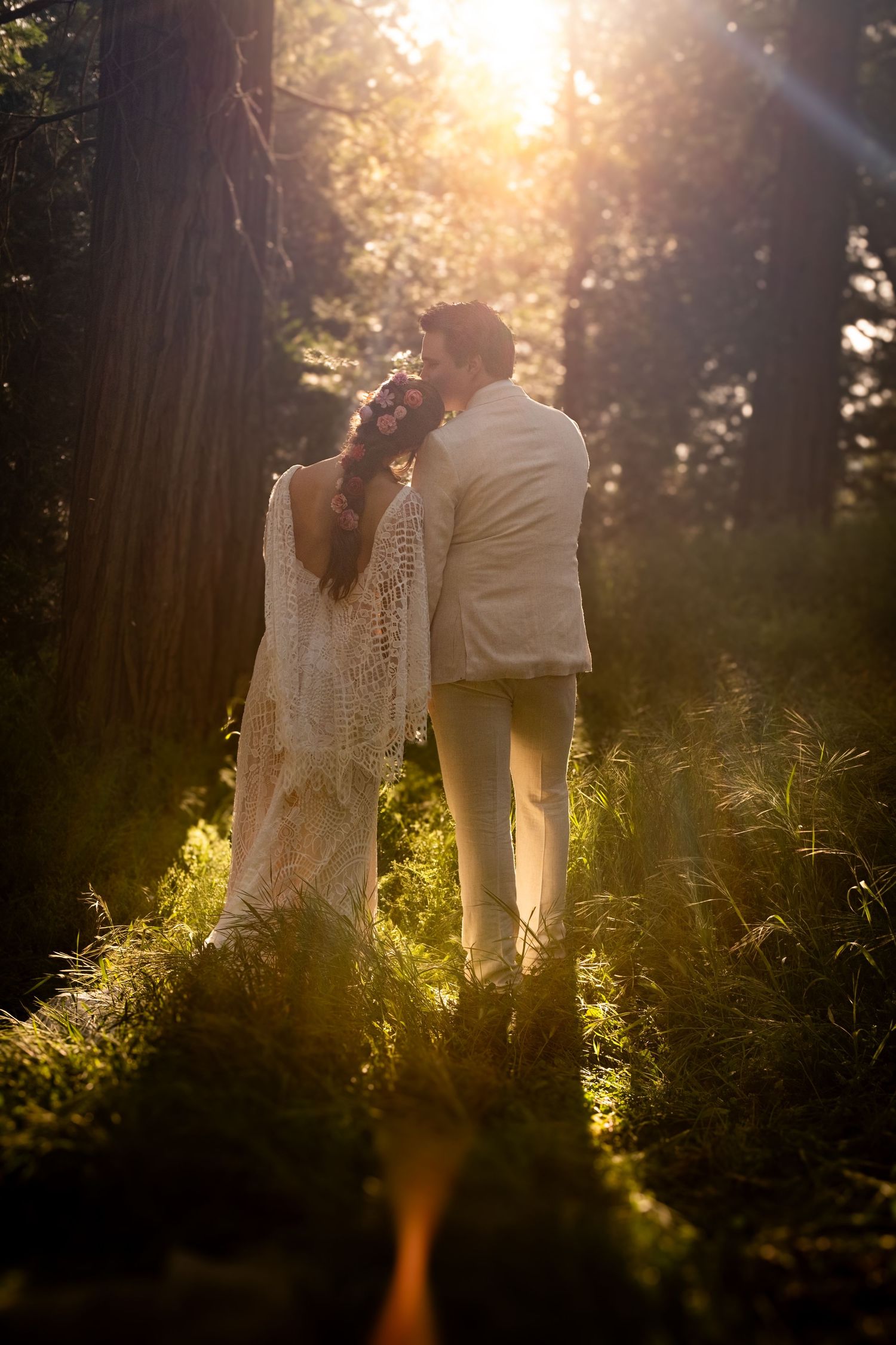 Bride and groom embracing in a sunlit forest, bride in lace gown, groom in white suit.