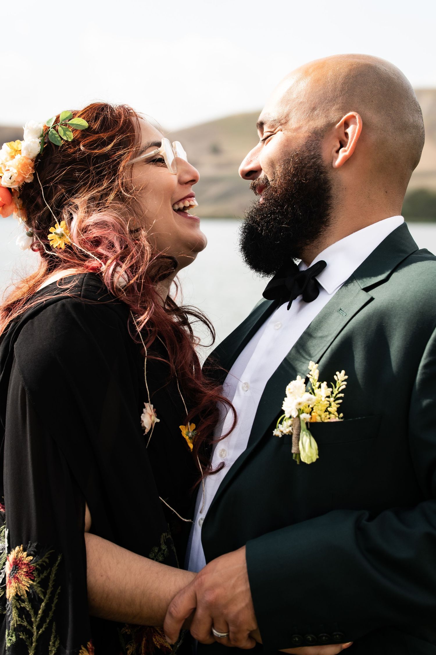 A joyful bride in black dress with floral crown and groom in dark tuxedo share a romantic moment outdoors.