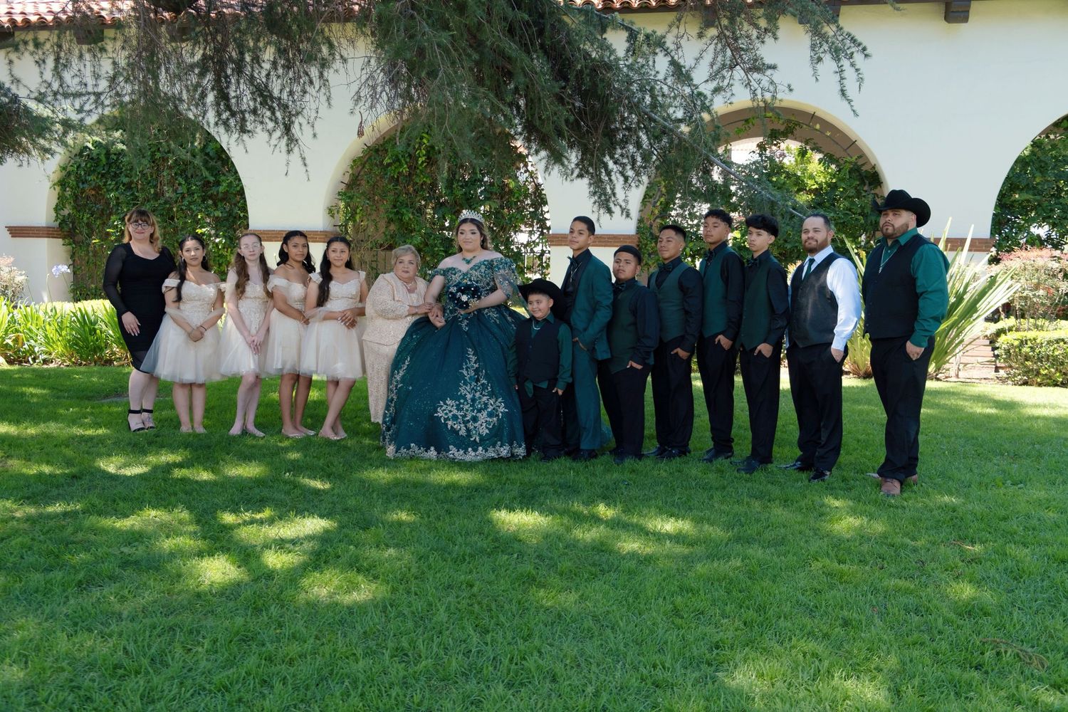Quinceañera court poses outdoors in green and blush formal attire on a sunny day.
