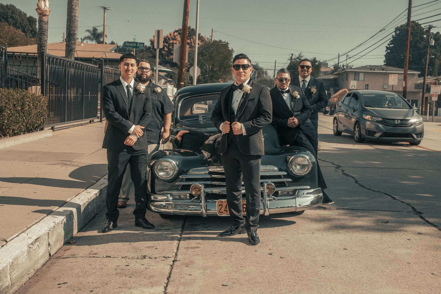 Groom and groomsmen posing confidently in dark suits beside a vintage black car on a sunny street.