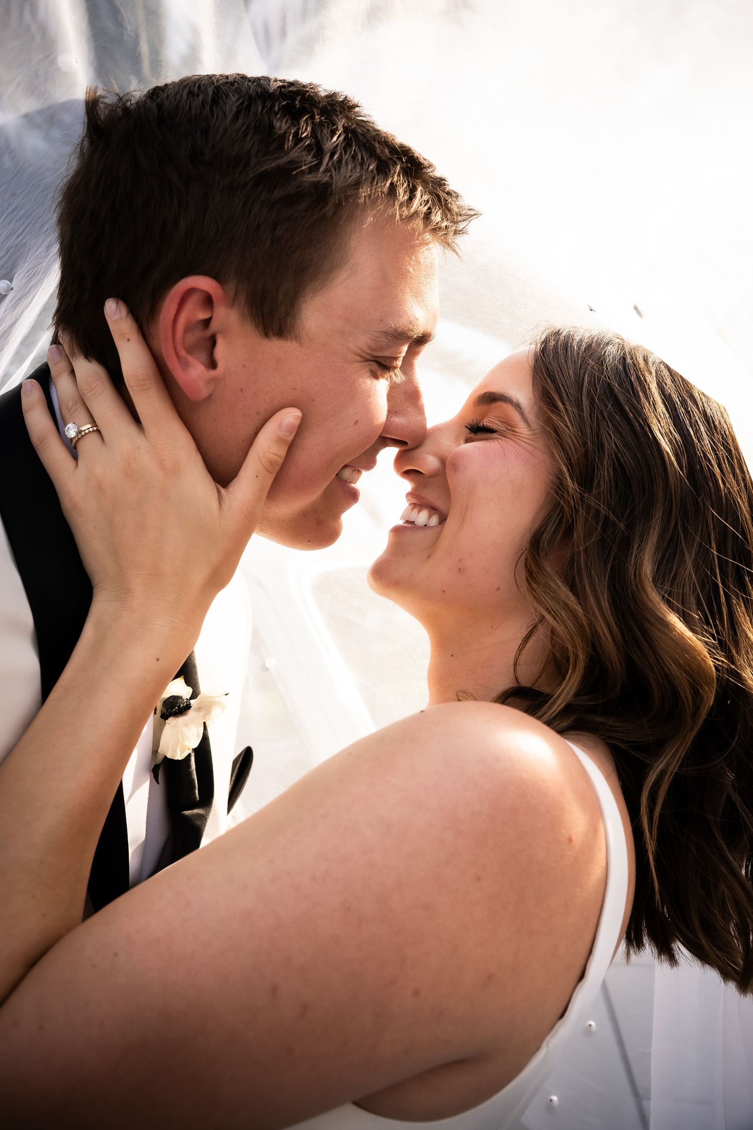 Smiling bride and groom nearly kissing under a veil in a joyful close-up wedding portrait.