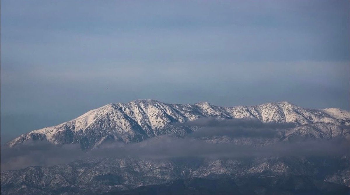 Snow-capped mountain range under a hazy blue sky, viewed from a distance with misty valleys below.