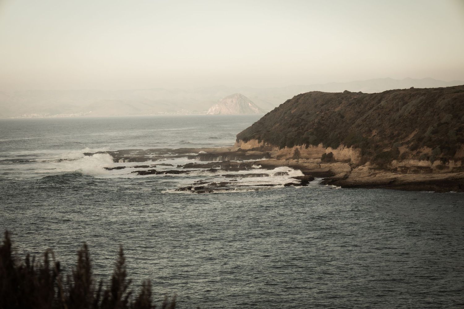 Rocky coastal cliffside with crashing waves, misty ocean horizon, and rugged shoreline landscape at dusk.