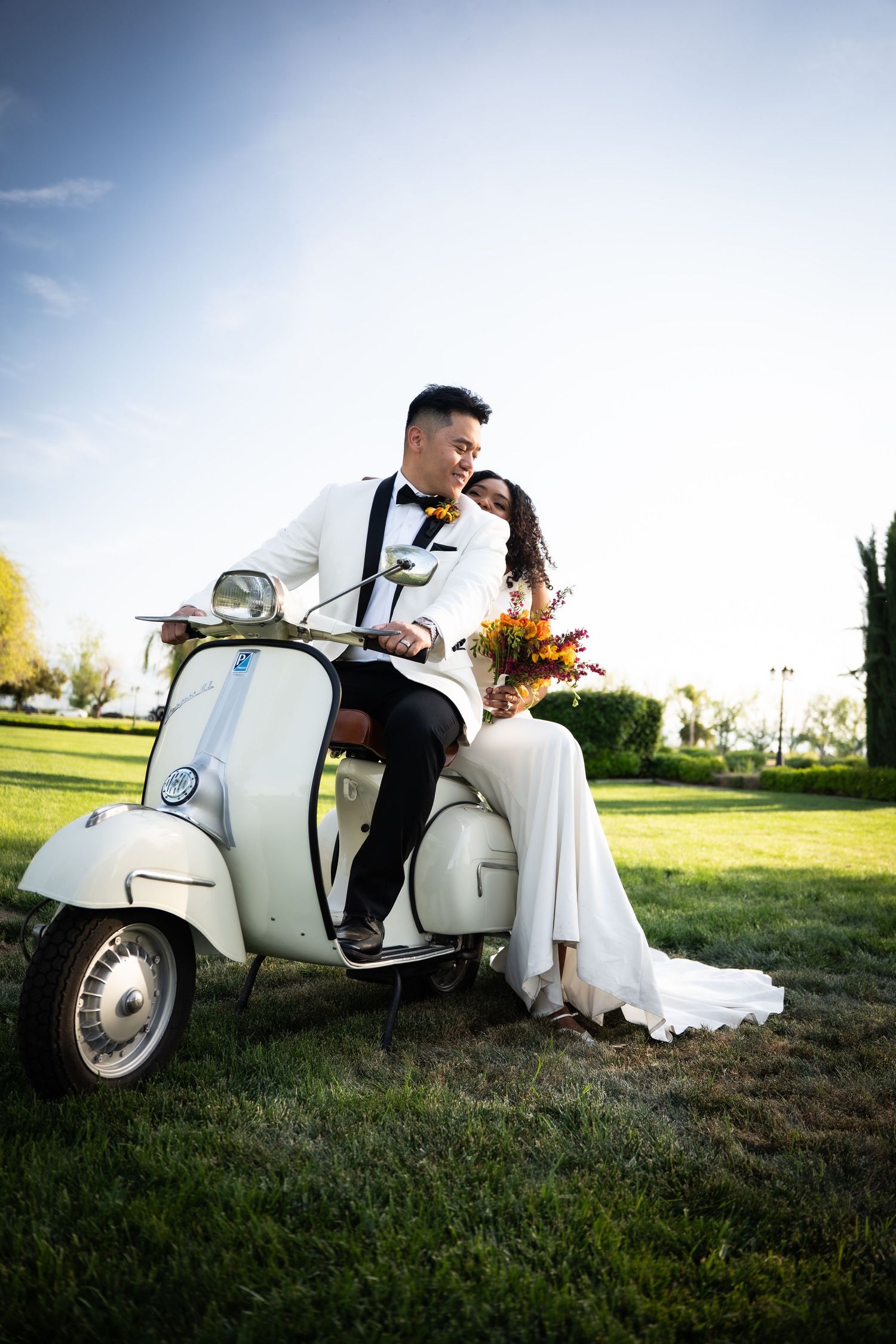 Newlywed couple sharing romantic moment on vintage white Vespa scooter in green park at golden hour.