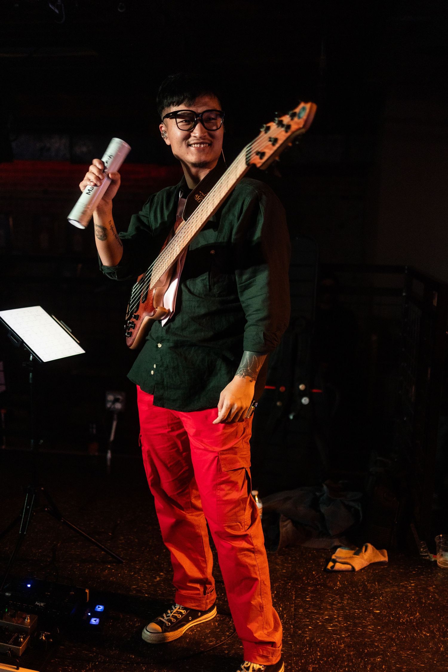 Smiling Asian bassist in red cargo pants and green shirt holding a can while posing with his bass guitar.