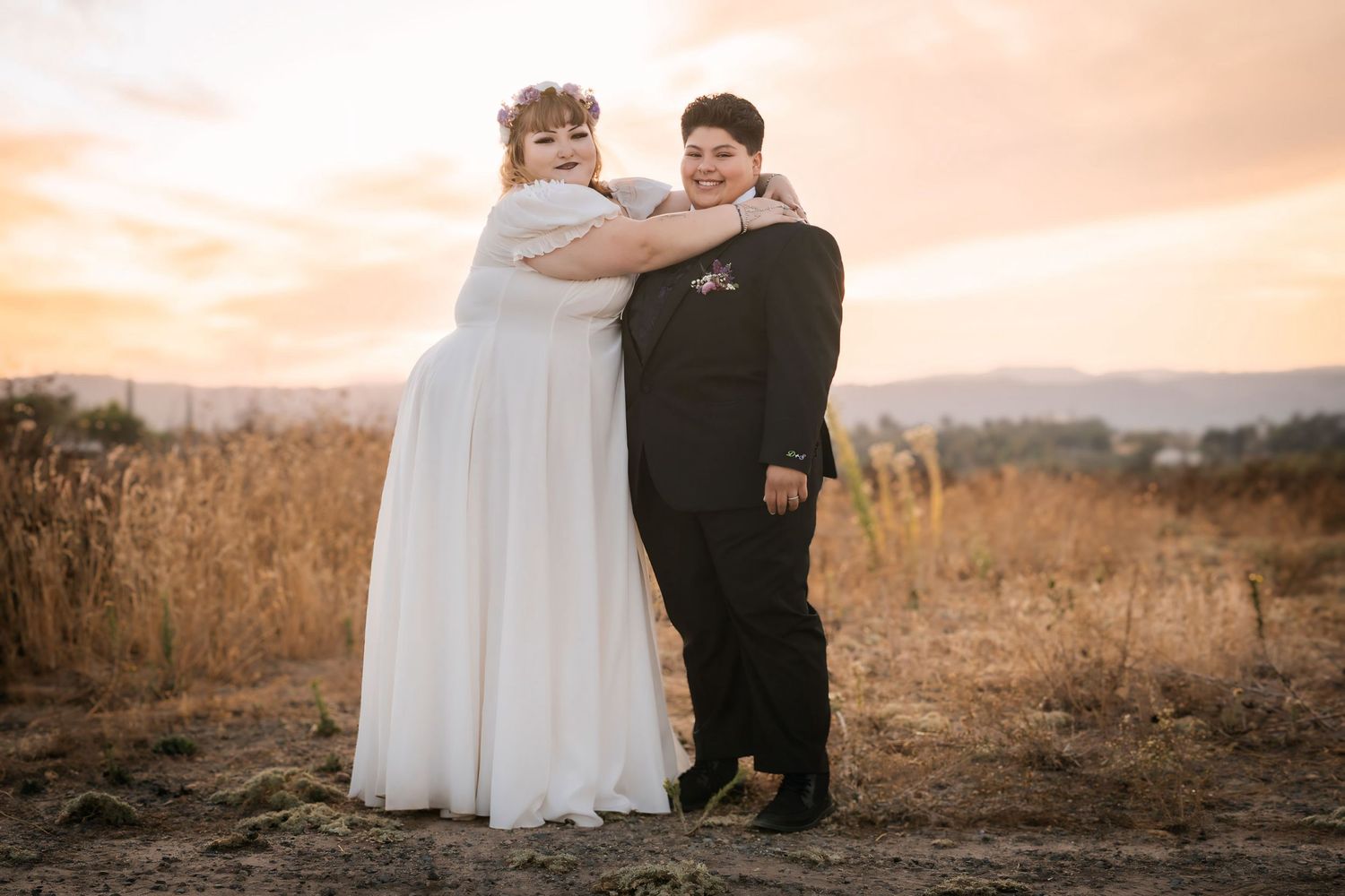 Two brides embrace at sunset in a golden field, one in a white gown and one in a black suit.