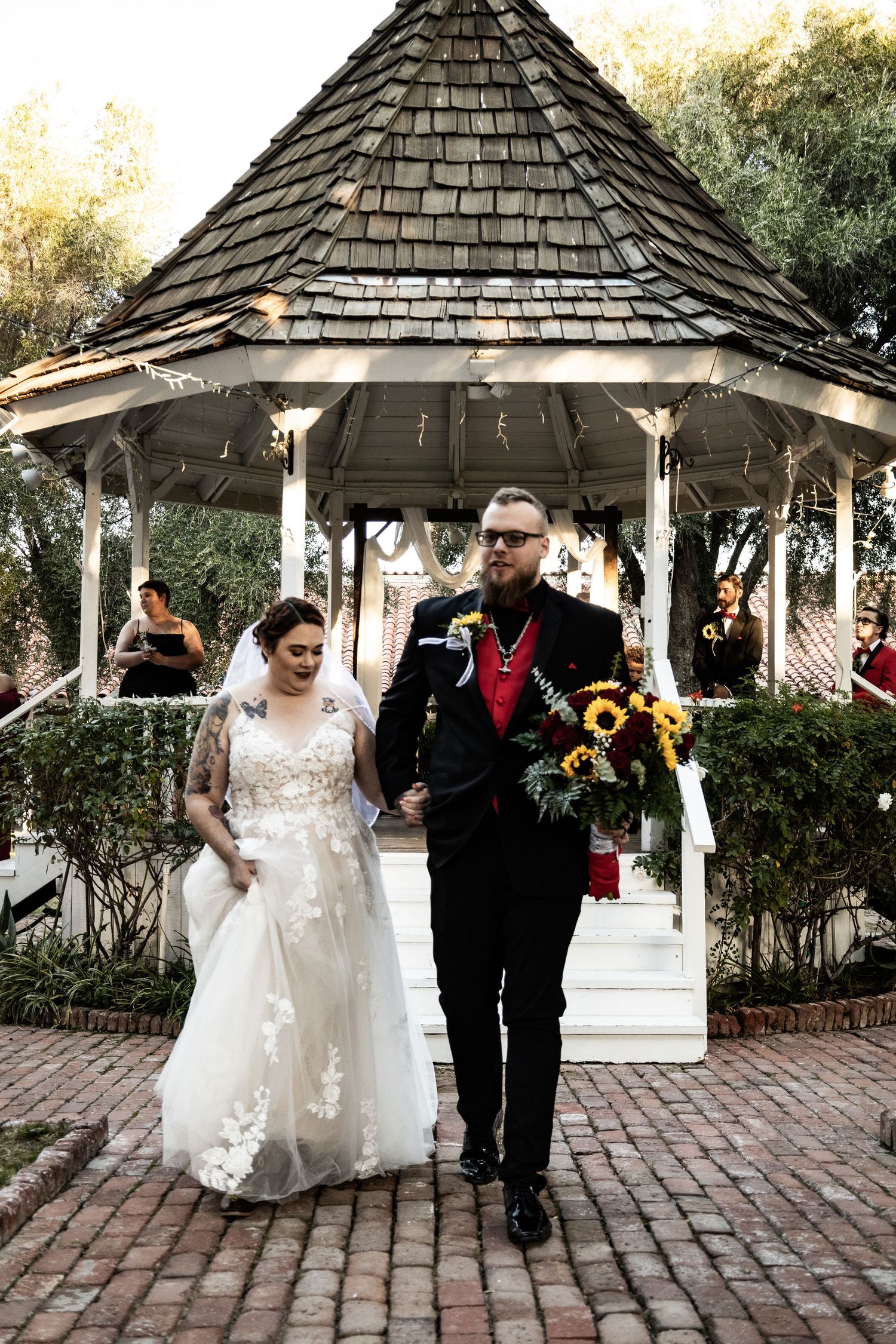 Bride and groom exiting a white gazebo, groom holding sunflower bouquet at outdoor wedding.