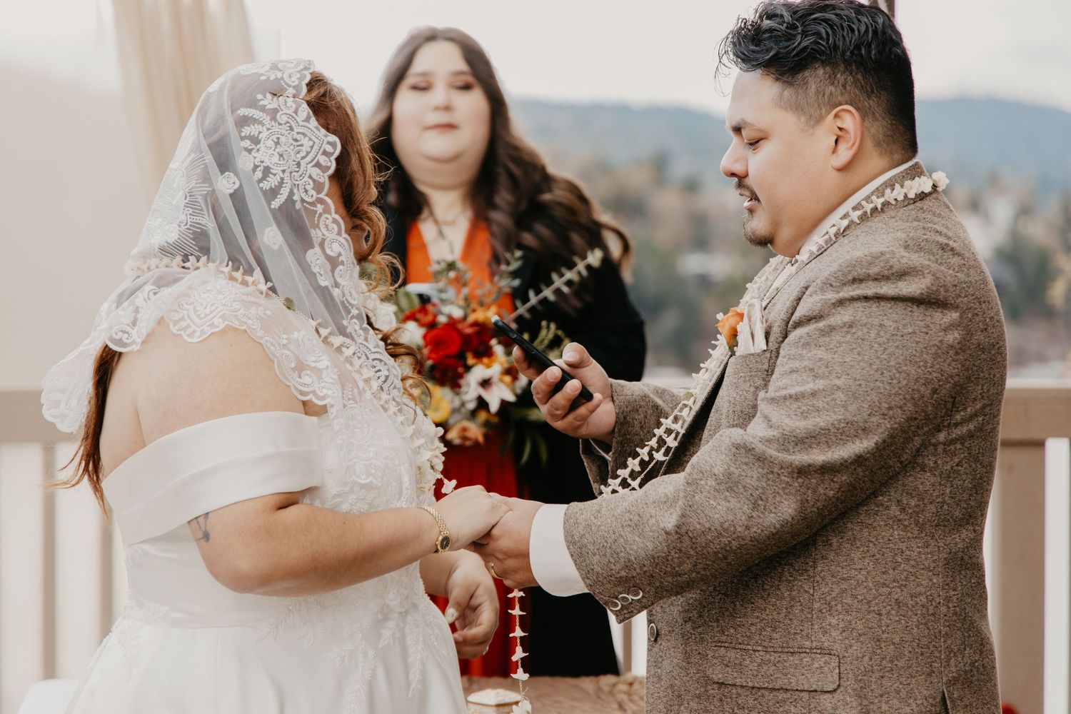 Groom places ring on bride's hand during outdoor wedding ceremony with officiant in background.