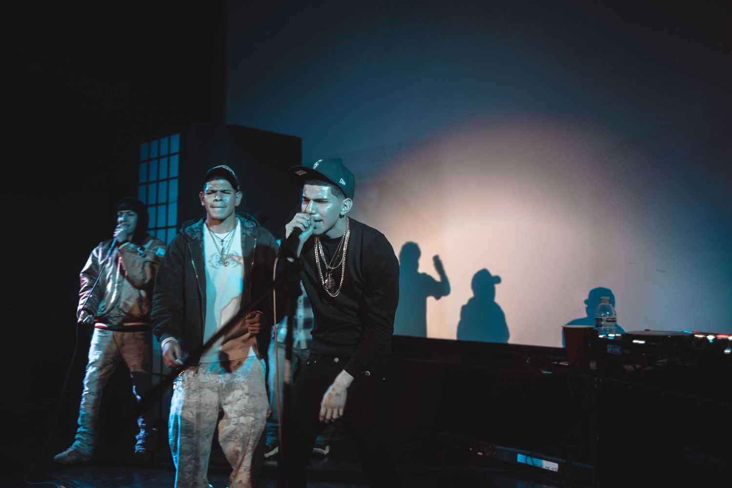 Three hip-hop artists performing on a dimly lit stage with dramatic shadows projected on the wall behind them.