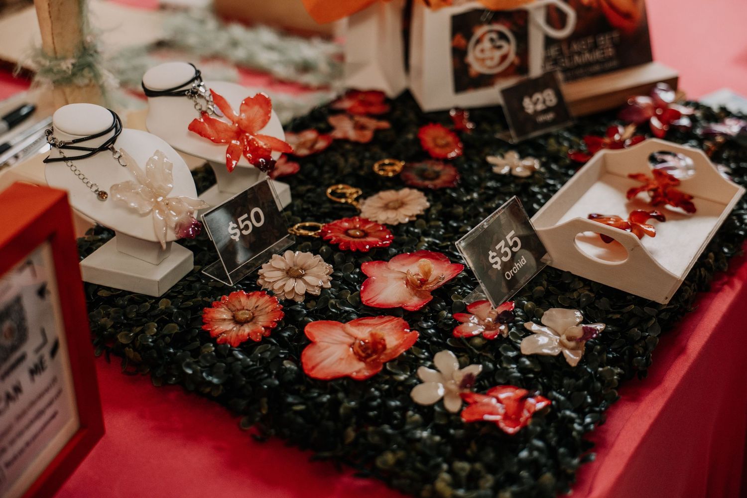 Handmade floral jewelry display with red flowers and price tags on a dark moss-covered table at a craft market.