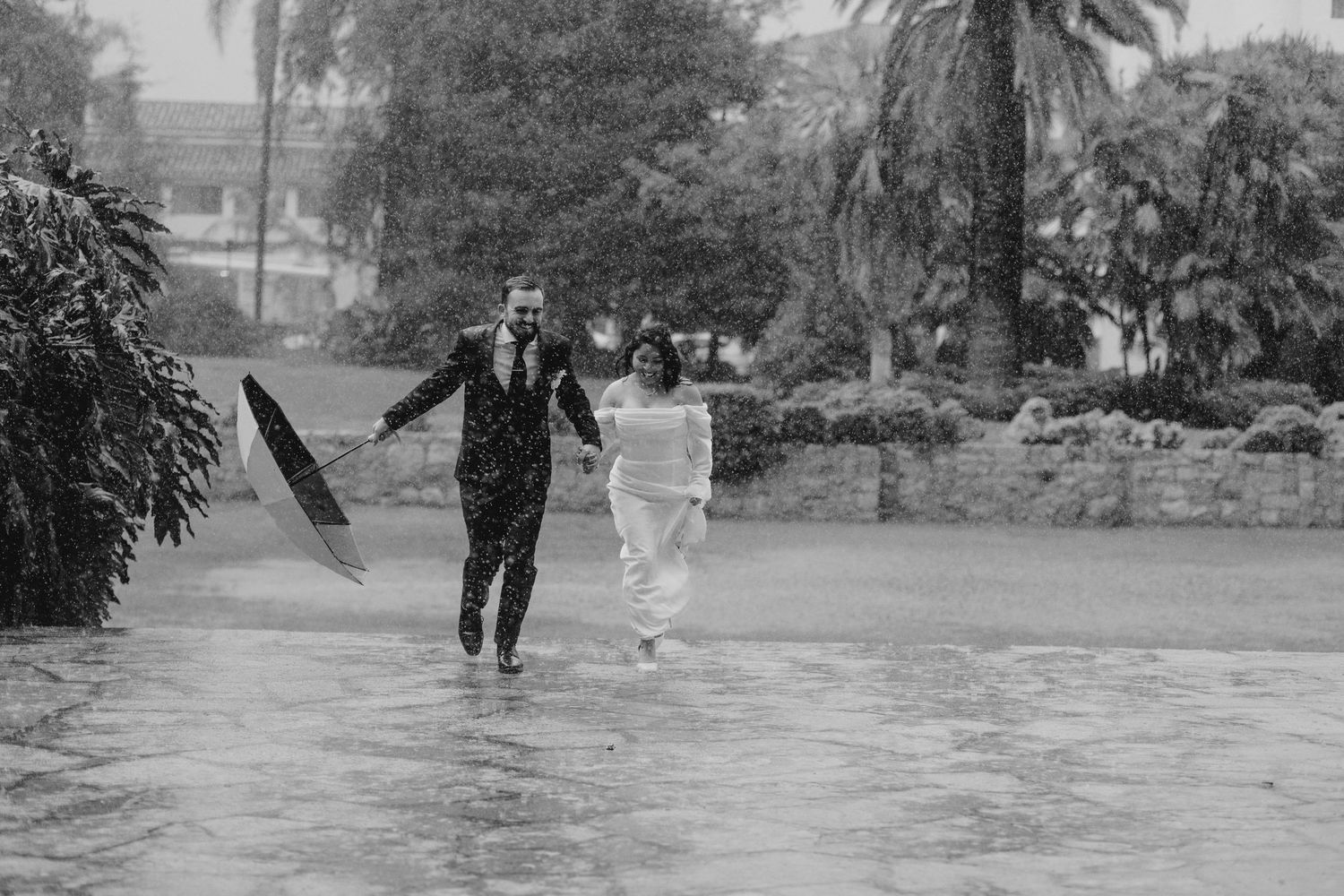 Black and white photo of couple running through heavy rain, groom holding umbrella at outdoor venue.