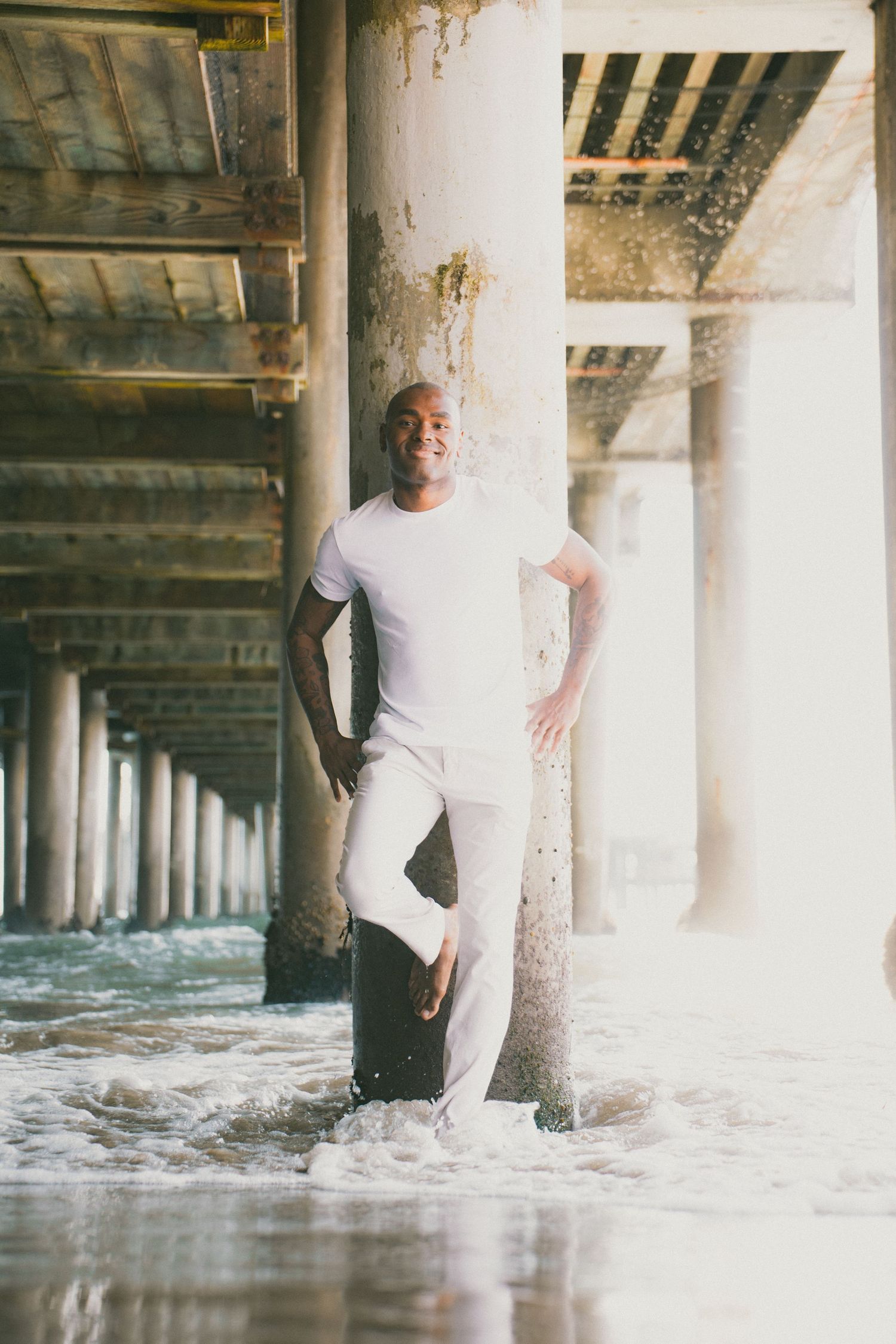 A smiling man in white outfit leans against a pier column as ocean waves splash around him.