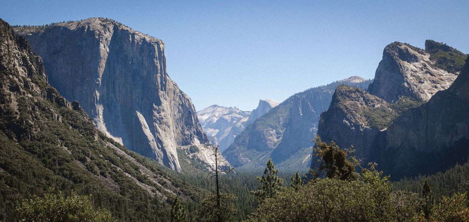 Panoramic view of Yosemite Valley with El Capitan, Half Dome, and lush green forests under a clear blue sky.