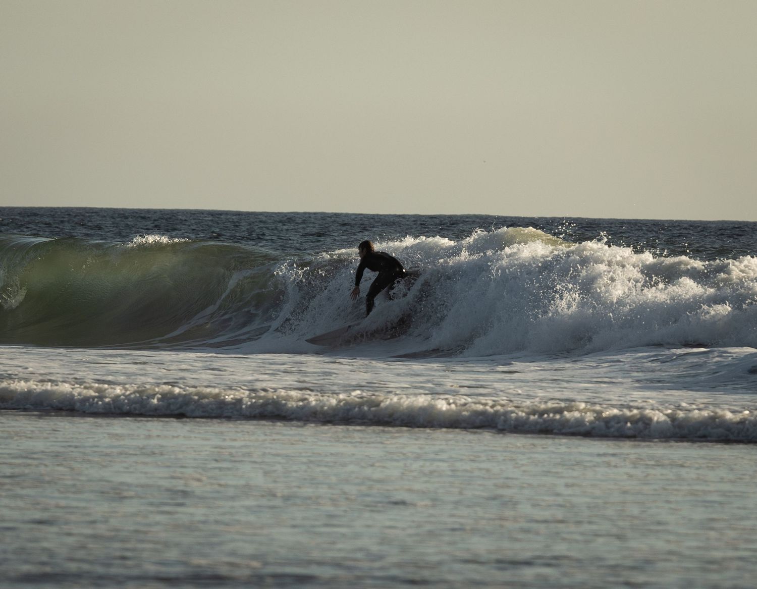 Surfer riding a wave in the ocean wearing a black wetsuit during golden hour light conditions.