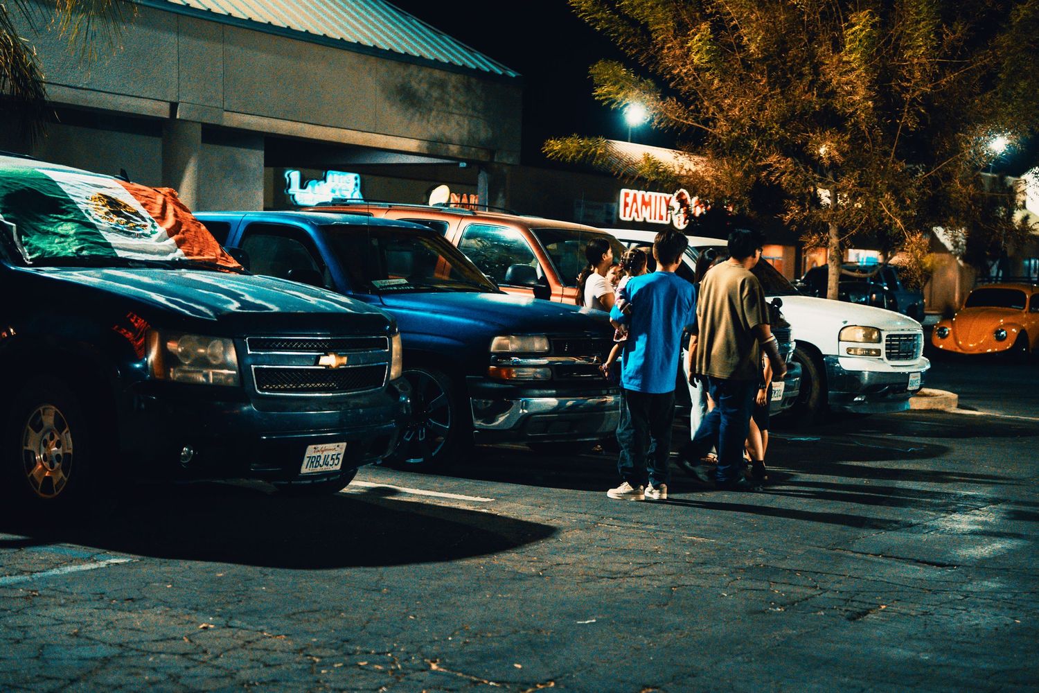 Group of people gathered around parked trucks in a parking lot at night outside a restaurant.