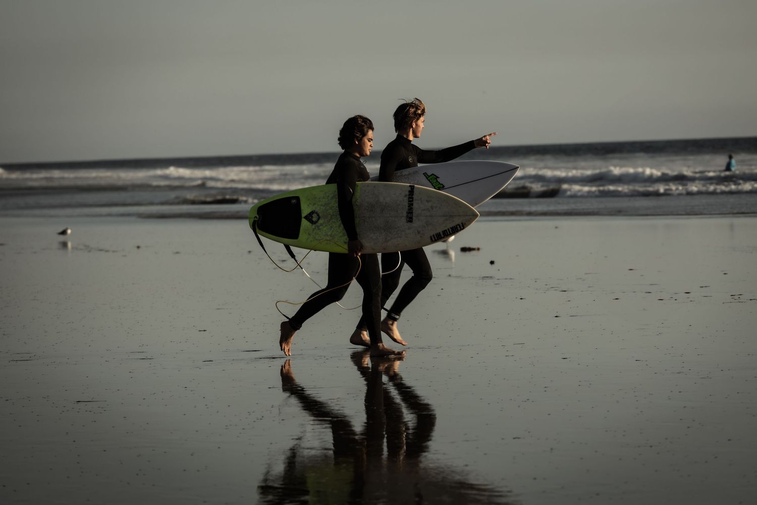 Two surfers in wetsuits carrying surfboards walking along a reflective wet beach at dusk.