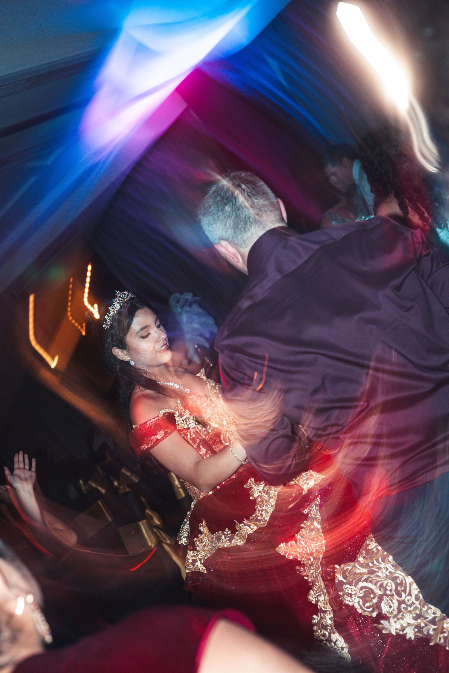 Dynamic motion blur photo of quinceañera dancer in red gown twirling under colorful lights.