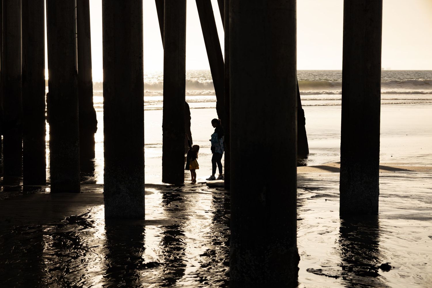 Silhouettes of two children standing under a pier with reflective wet sand and ocean waves behind them.