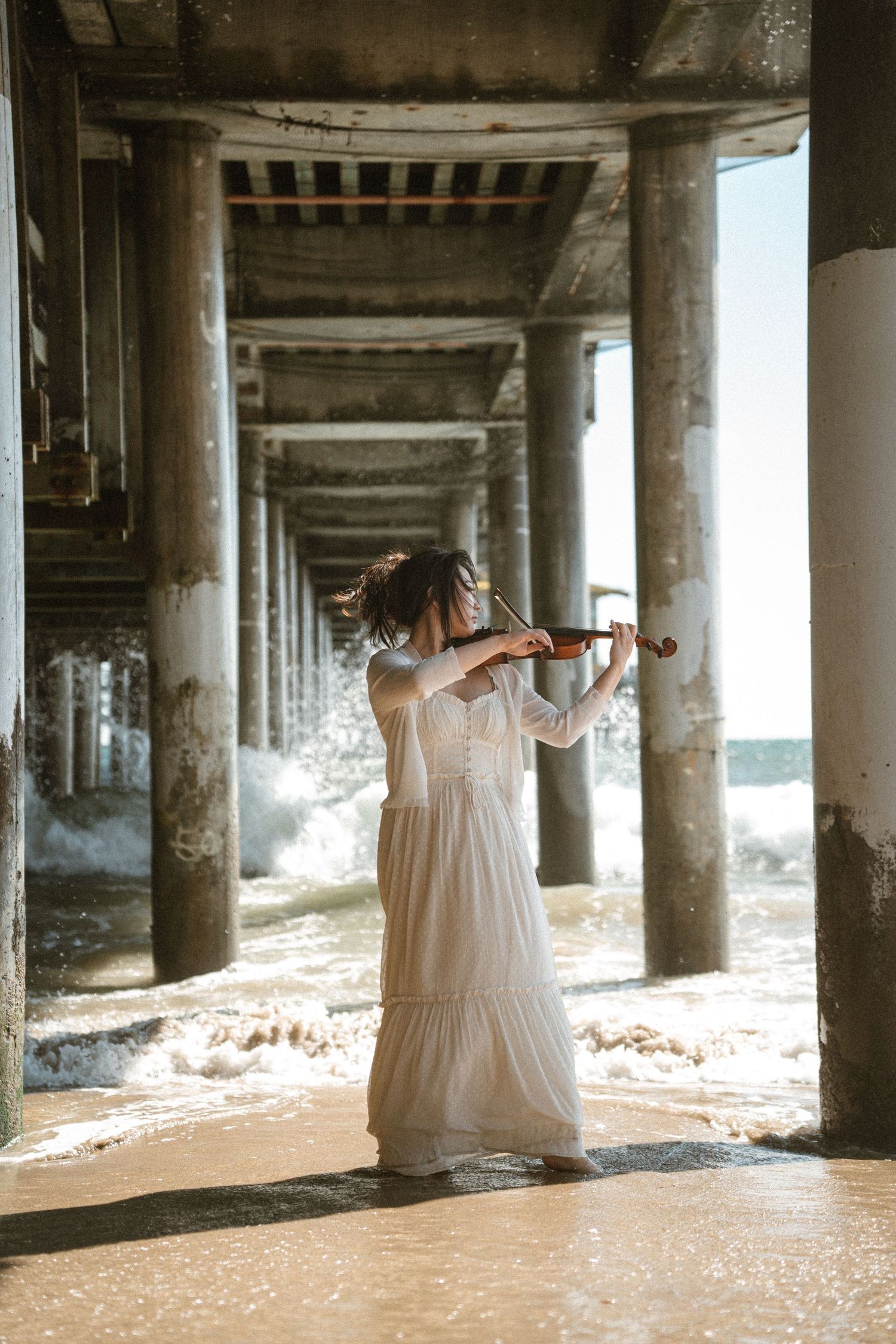 Elegant woman in white gown playing violin under a seaside pier.