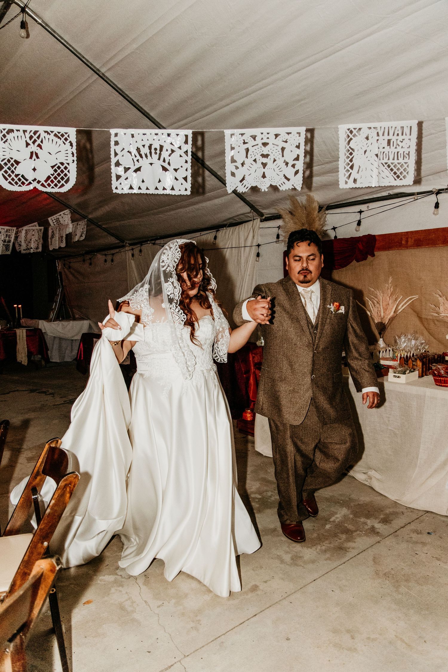Bride in lace gown and groom in tweed suit enter reception tent with papel picado decor.