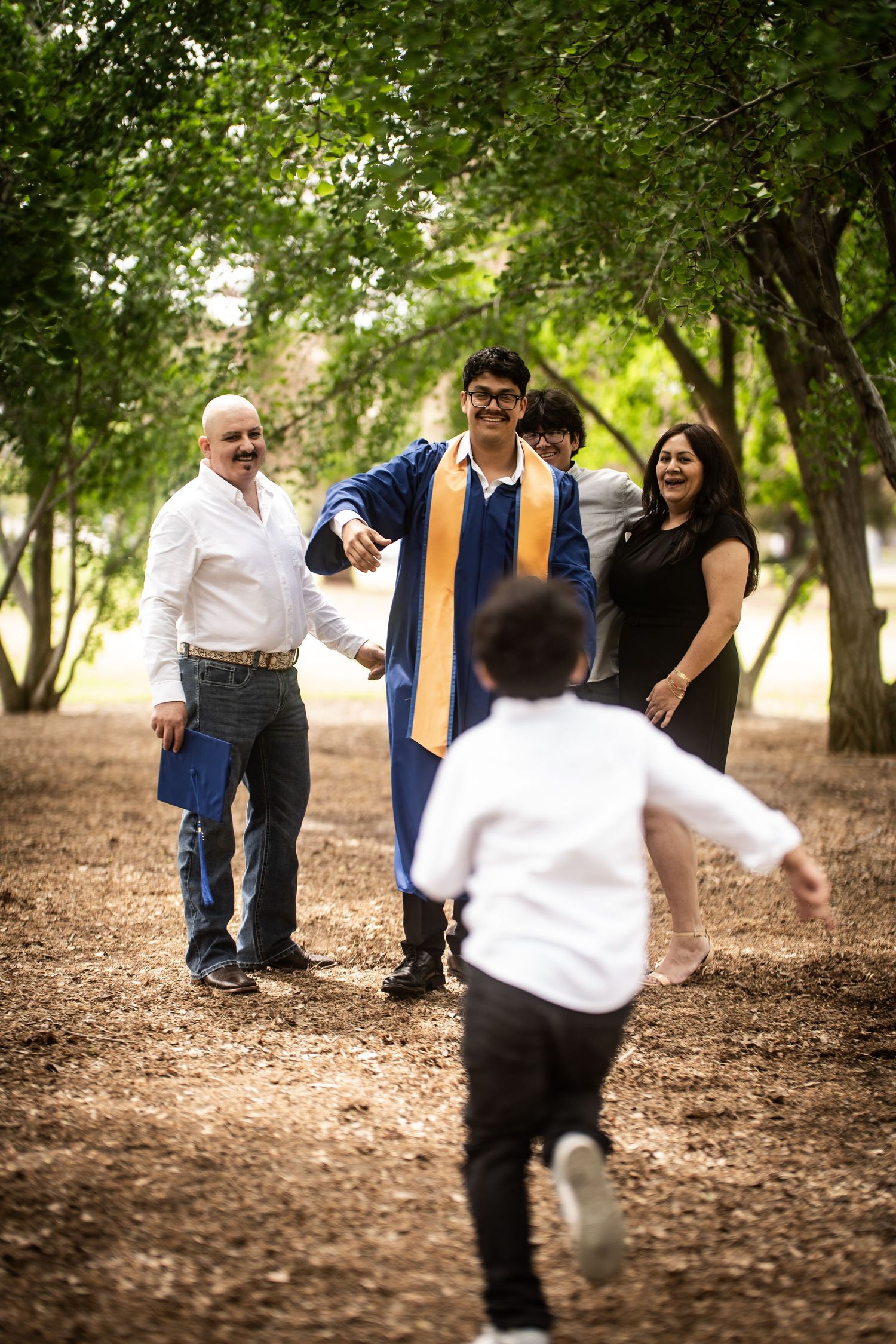 A joyful graduate in blue gown celebrates with family as a child runs toward them outdoors.
