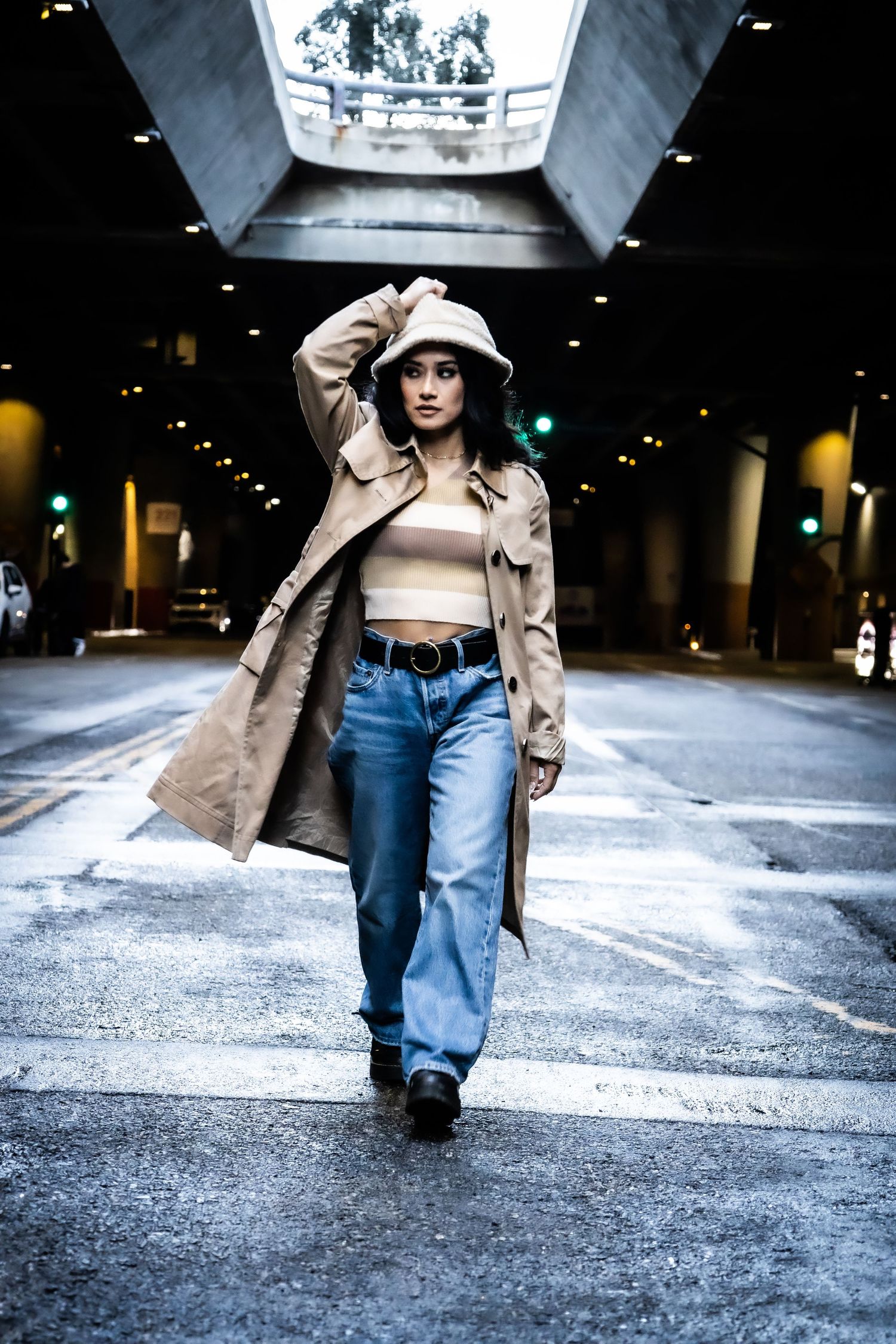 A woman in a trench coat and bucket hat walking under an urban overpass at night.