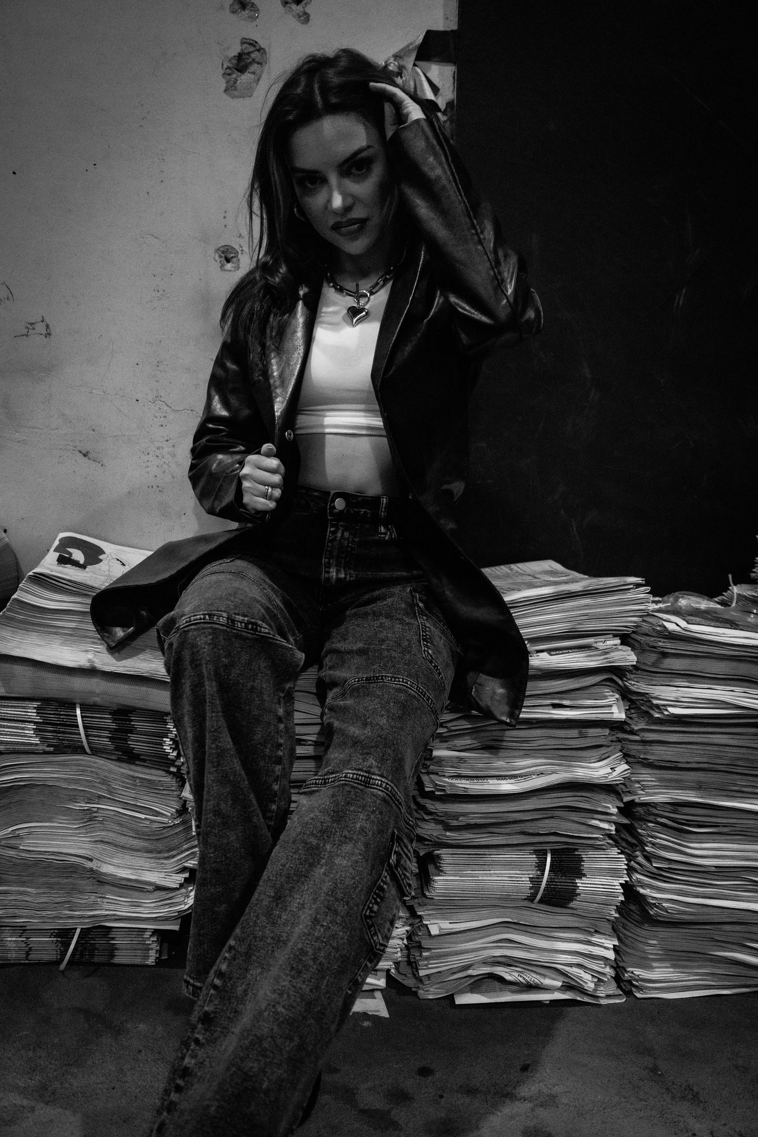 A woman in a leather jacket sitting on stacked newspapers in a gritty black and white photo.