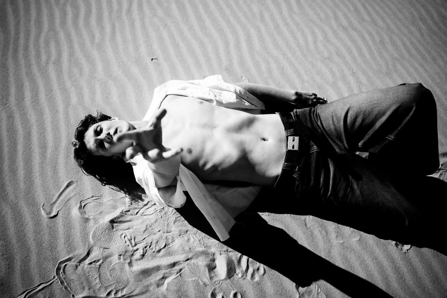 Black and white photo of a shirtless man lying on desert sand dunes with an open shirt.