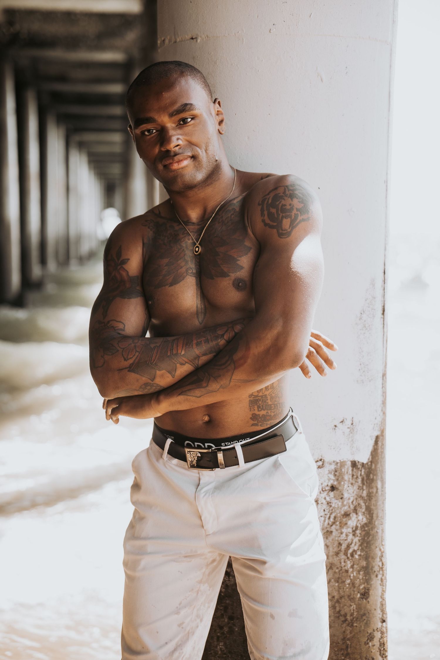 A tattooed shirtless man with crossed arms poses confidently under a pier at the beach.