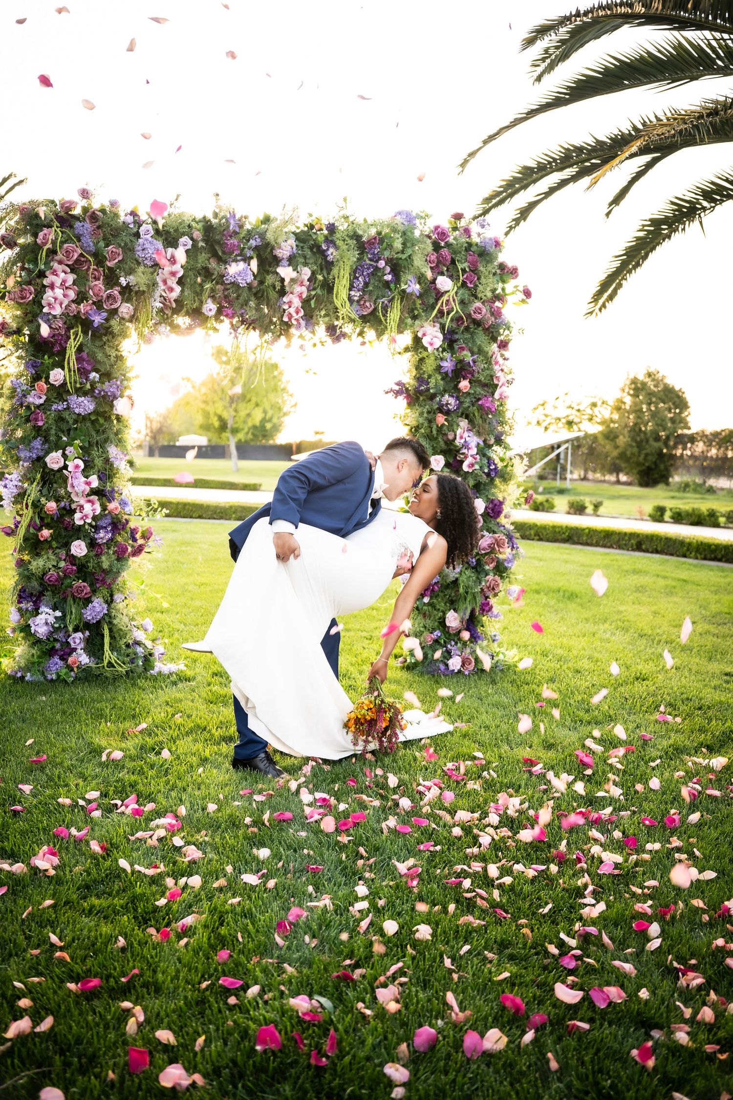 A couple shares a dip kiss under a floral arch with rose petals falling around them.