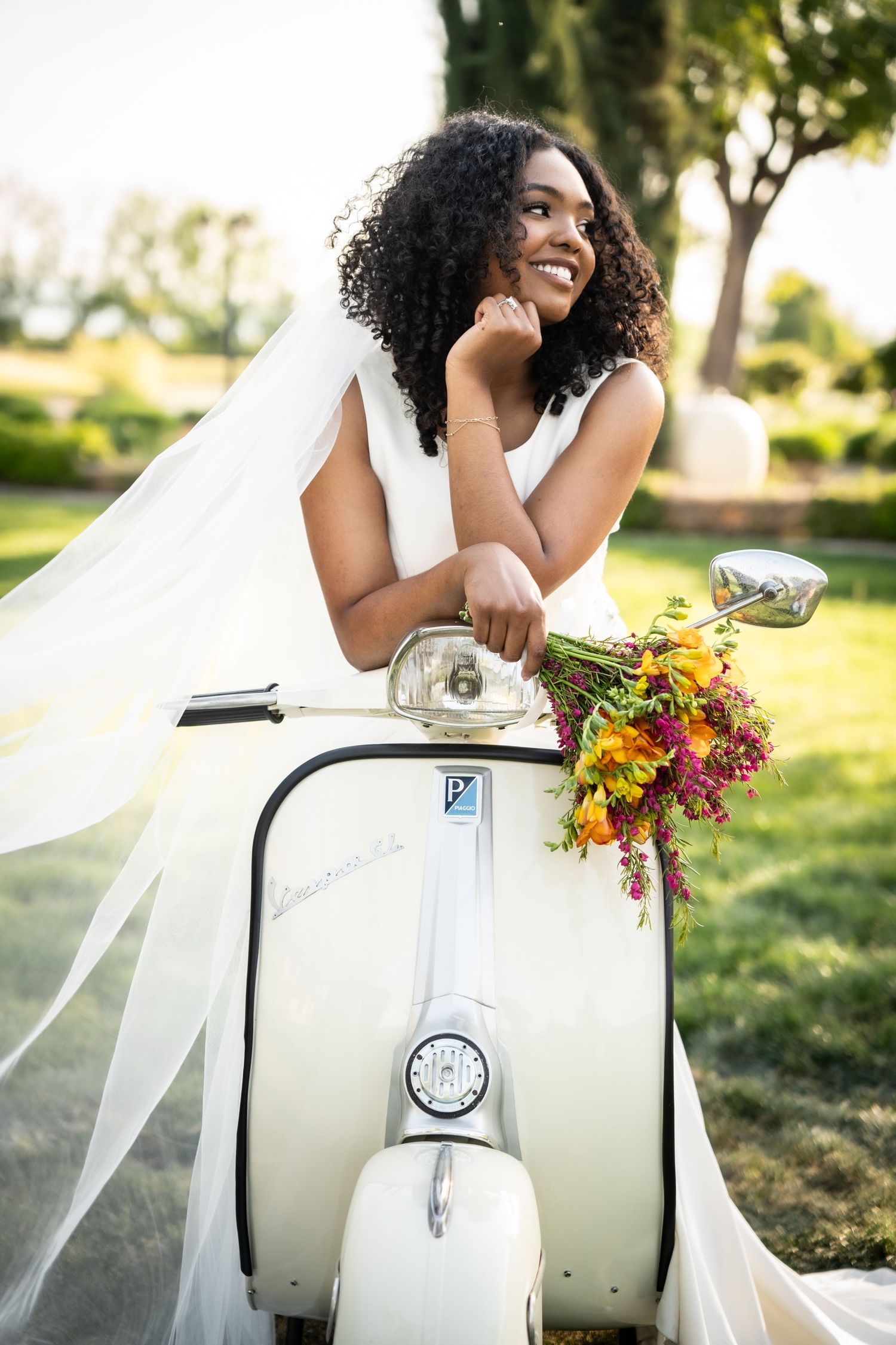 Smiling bride in white dress and veil posing with a vintage cream Vespa scooter holding colorful bouquet outdoors.