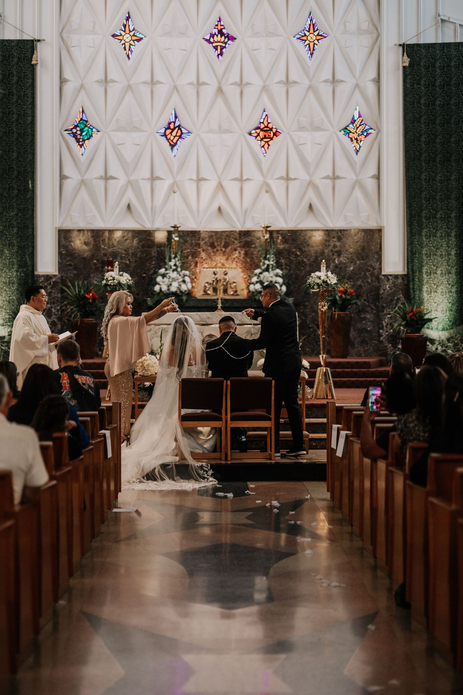 Bride and groom during a traditional lasso ceremony in a modern Catholic church with stained glass windows.