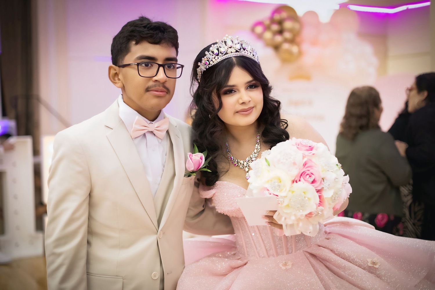 Quinceañera girl in pink ball gown and tiara poses with escort holding floral bouquet.