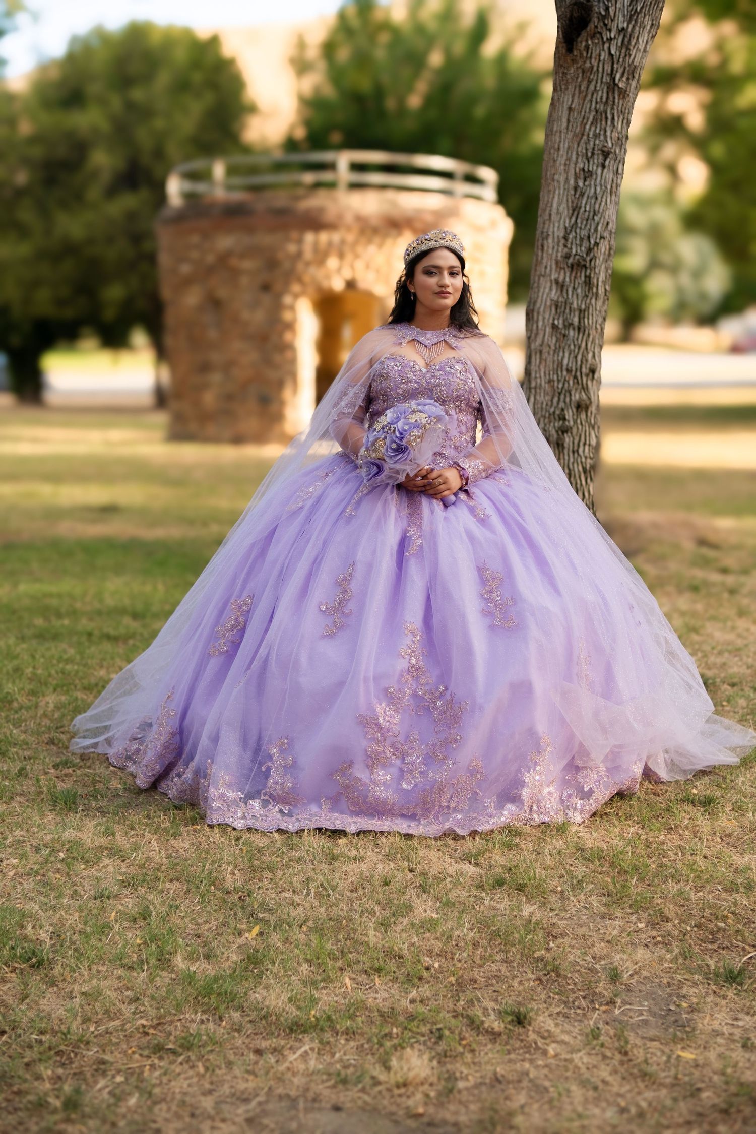 Young woman in lavender quinceañera ball gown with cape holds bouquet in park setting.