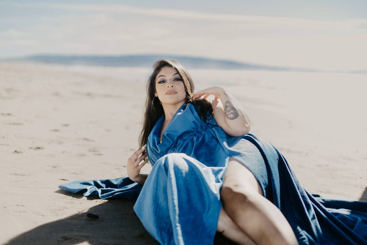 Woman in flowing blue gown reclines on sandy beach with ocean in background.