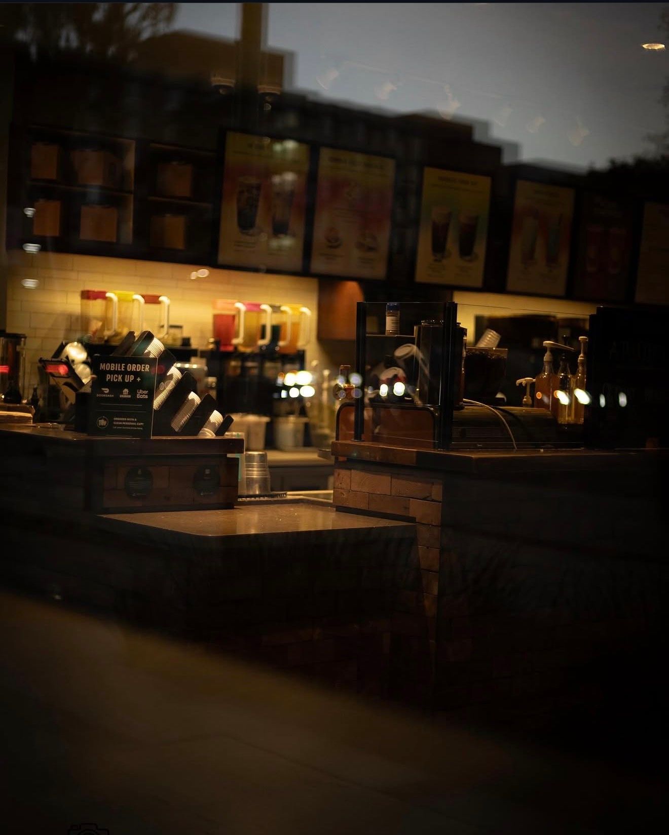 Dimly lit Starbucks interior viewed through glass, showing menu boards and counter at night.