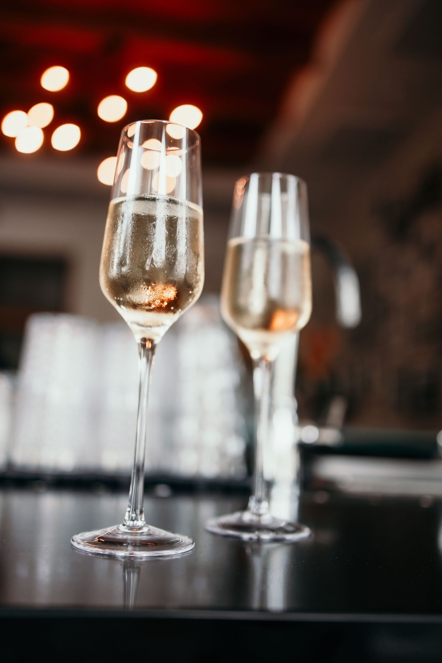Two elegant champagne flutes filled with sparkling wine on a dark bar surface with bokeh lights in background.