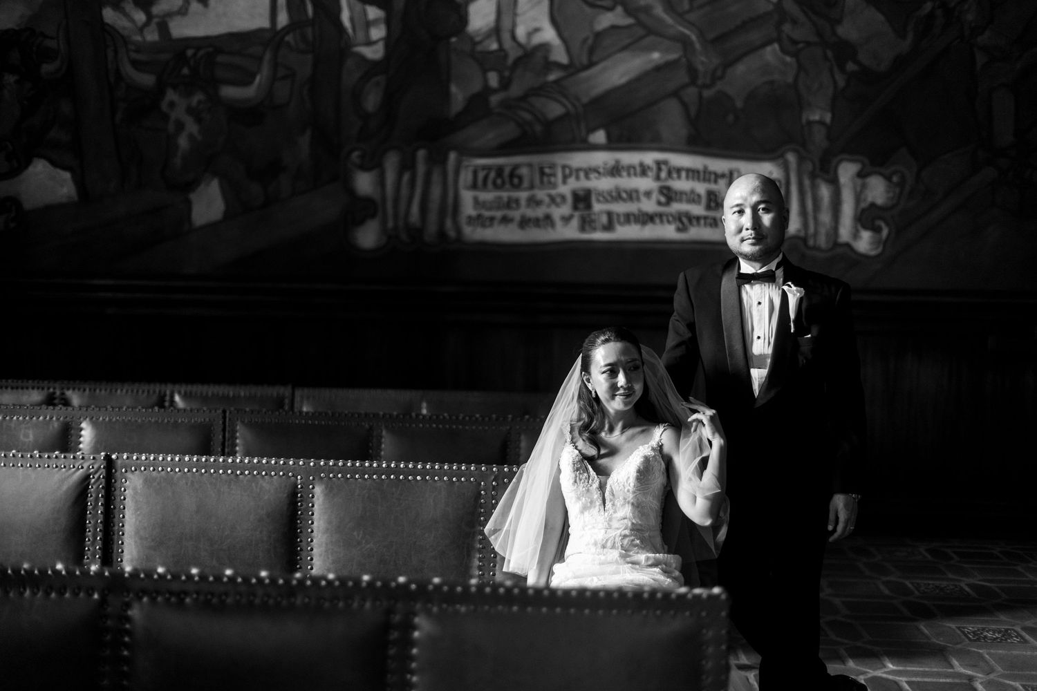 Black and white wedding photo of bride and groom standing in an ornate hall with murals.
