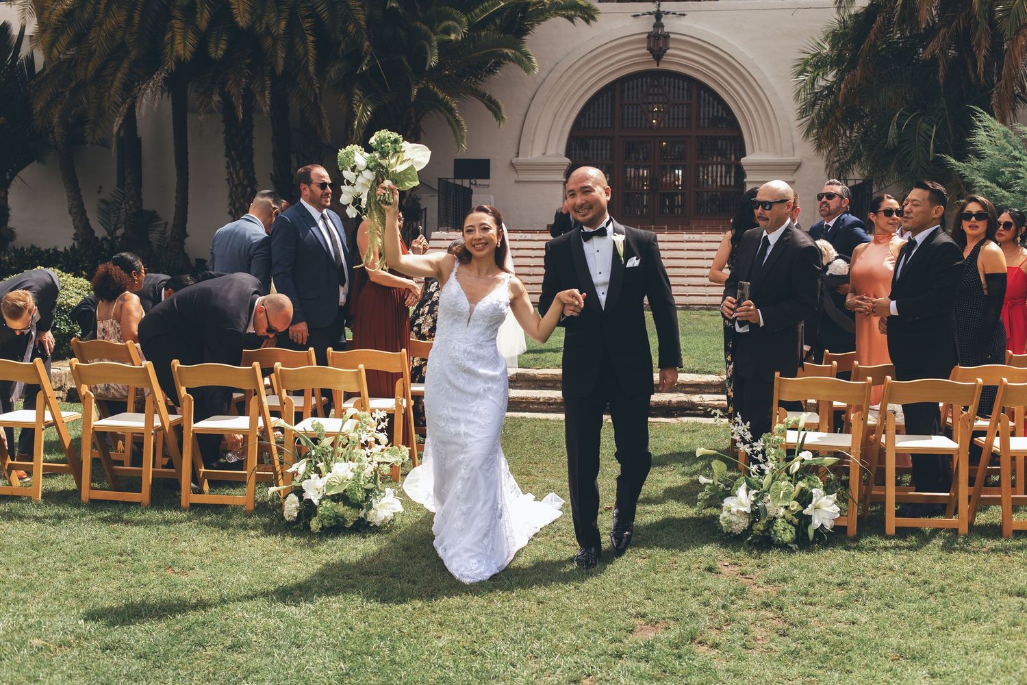 Bride and groom walk down outdoor wedding aisle, surrounded by guests and floral arrangements, at a sunlit venue.