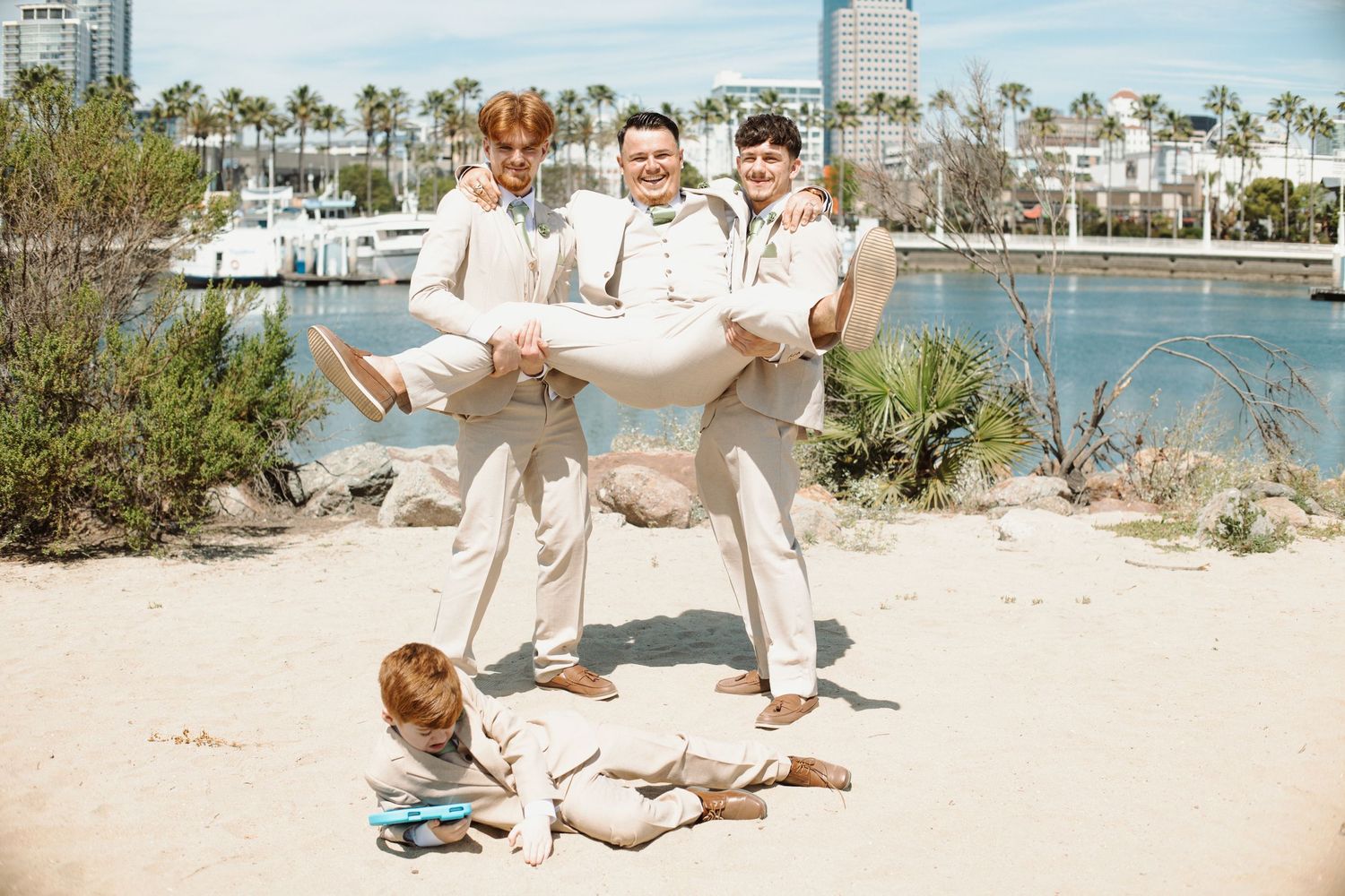 Groomsmen in beige suits playfully lift groom on sandy beach with waterfront backdrop at outdoor wedding.